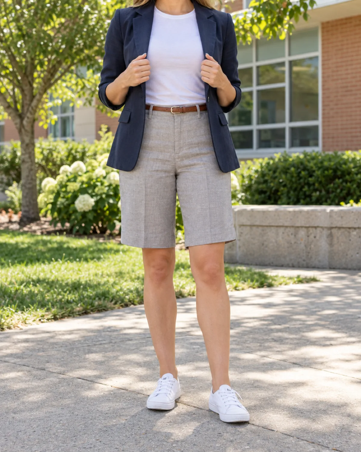A woman in a school courtyard wearing stone-grey linen bermuda shorts with a navy cotton blazer held open to show a white tee underneath, a brown belt and white sneakers, school building behind her