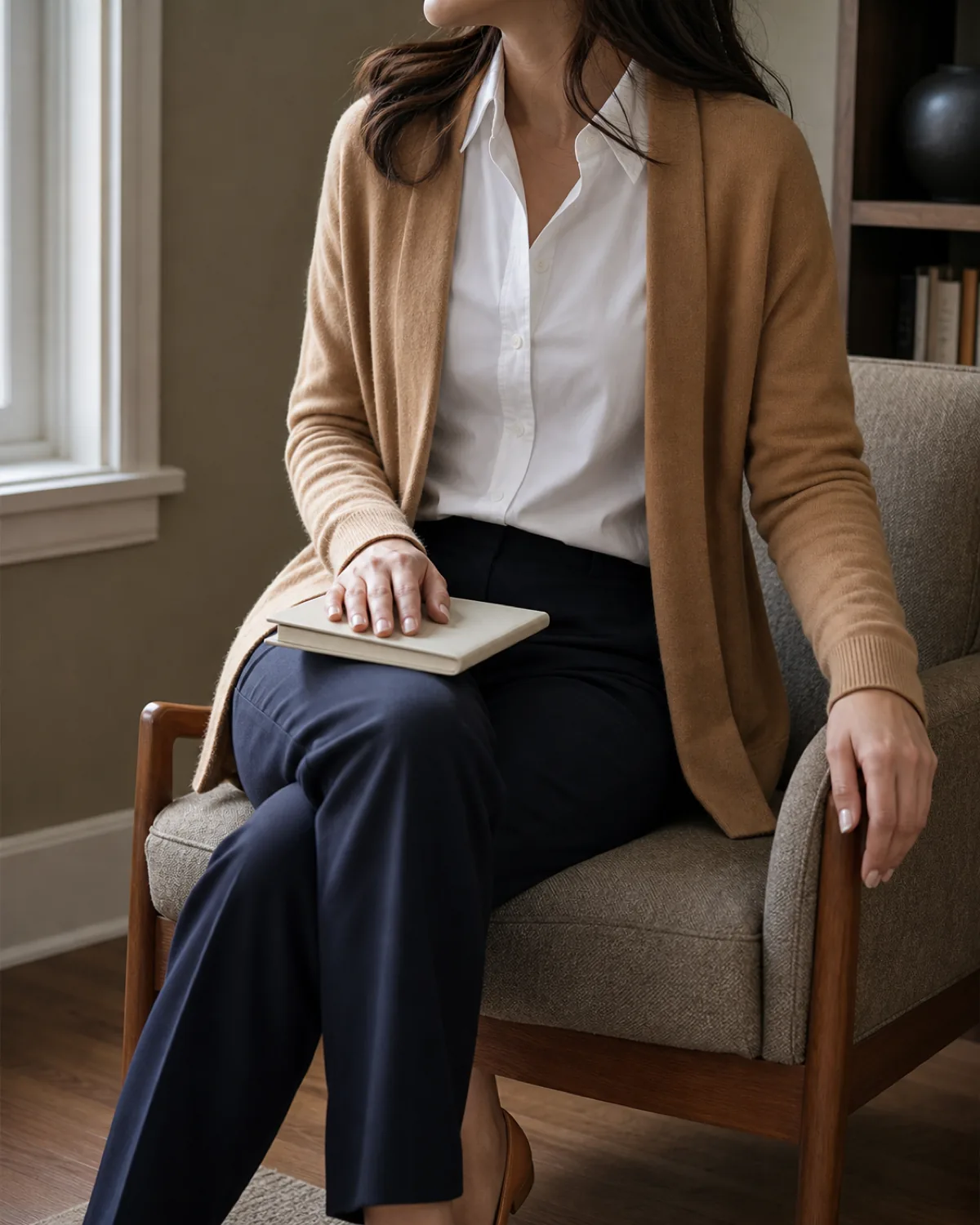 A woman seated in a mid-century armchair with legs crossed, wearing a camel cardigan open over a white blouse and dark navy trousers, one hand resting on a notebook in her lap