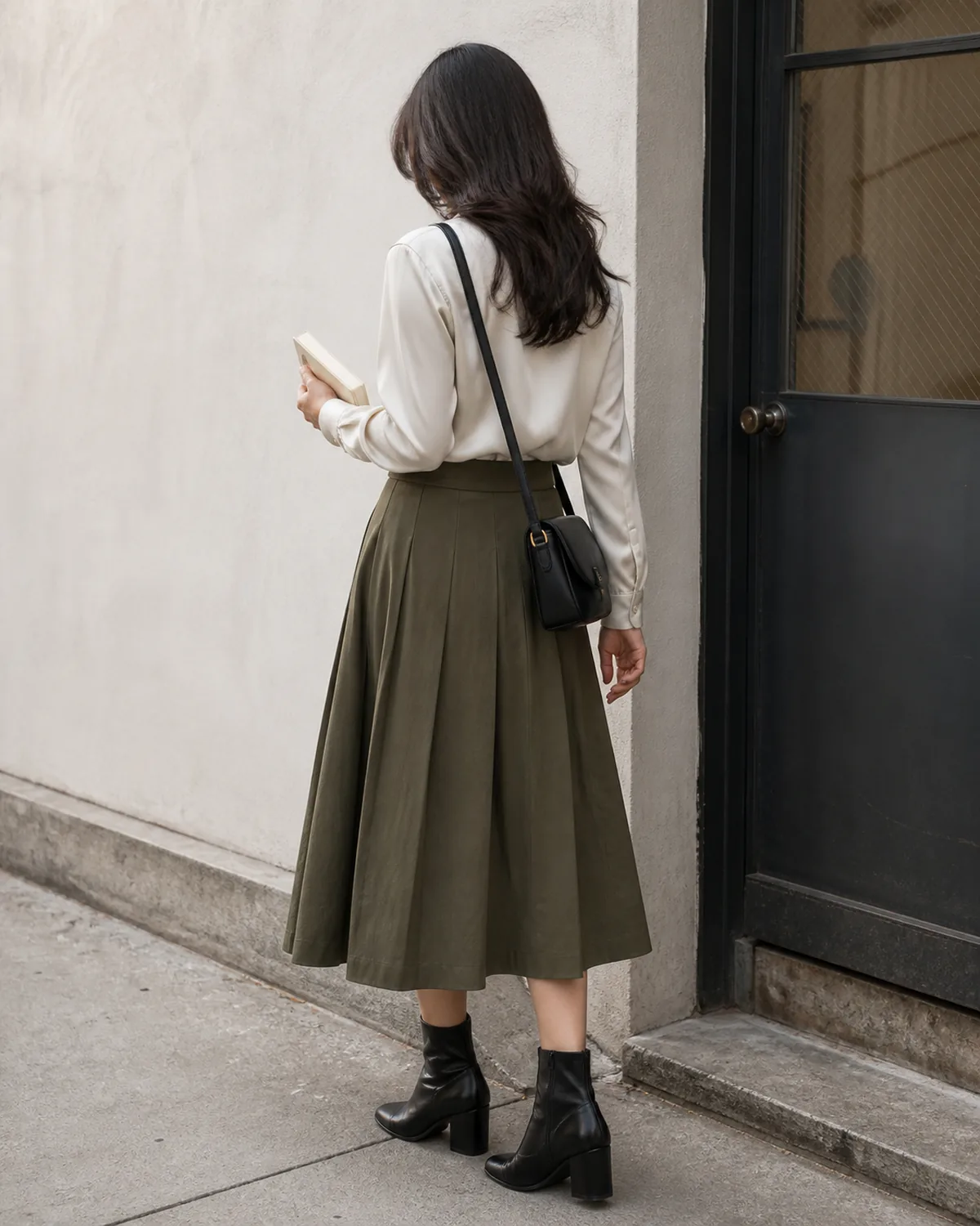 A woman seen from behind in an ivory blouse tucked into an olive pleated midi skirt with black ankle boots and a small crossbody bag, standing near a metal-frame door