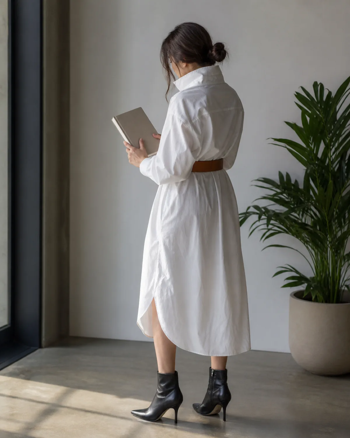 A woman in a back-three-quarter view looking down at a book, wearing an oversized white button-down shirt belted at the waist with a wide cognac belt creating a shirtdress silhouette that falls below the knee, with black pointed-toe ankle boots and a potted plant nearby