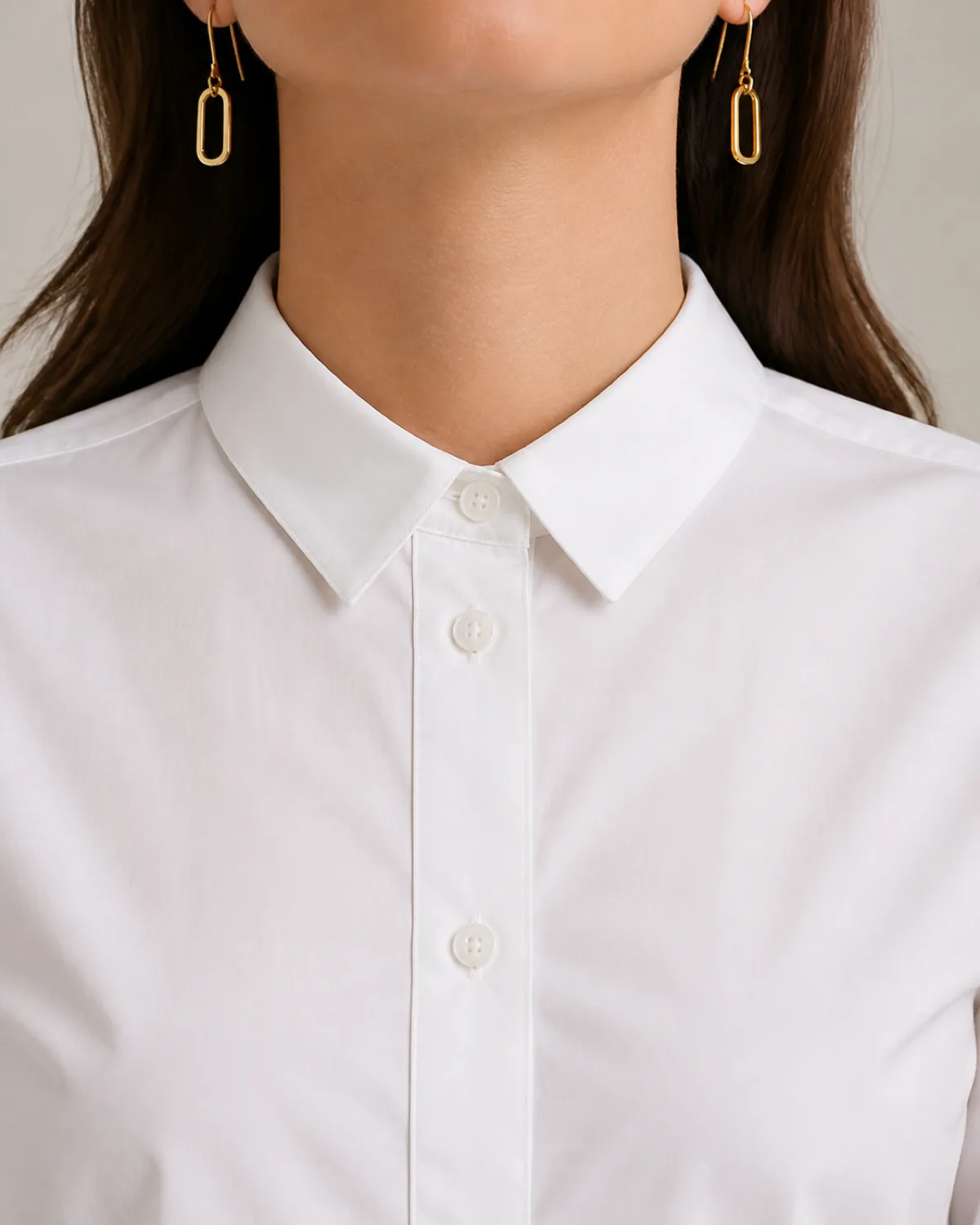 Close crop on shoulder and collar showing a white button-down shirt buttoned all the way to the top with simple gold geometric drop earrings, the collar sitting snug and the button placket aligned