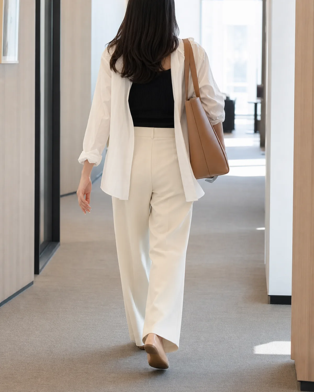 A woman walking away from camera in an office hallway wearing an unbuttoned white button-down shirt open over a fitted black ribbed tank top with cream wide-leg trousers, tan leather tote on her shoulder, and nude heeled shoes