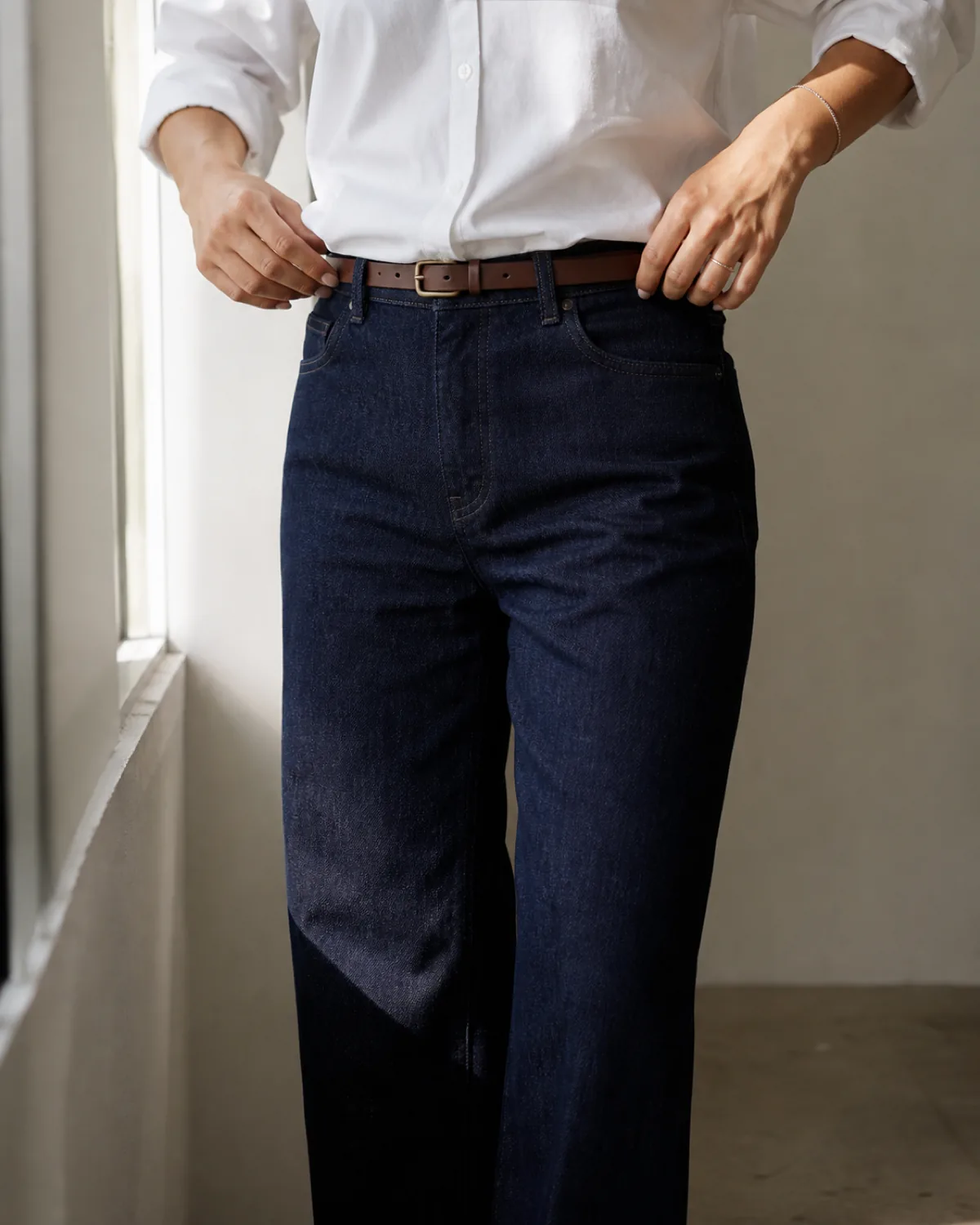 Close crop of dark indigo straight-leg jeans with a white button-down shirt tucked in and a cognac leather belt at the waist