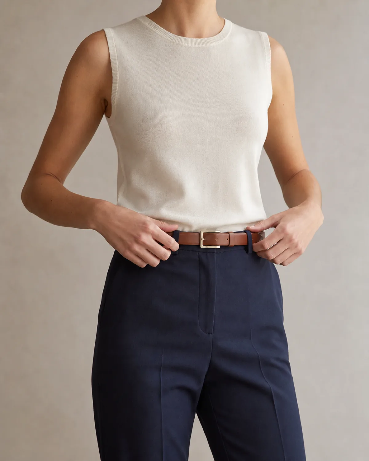 A woman wearing an ivory fine-gauge knit shell tucked into navy trousers with a cognac leather belt in a mid-torso crop
