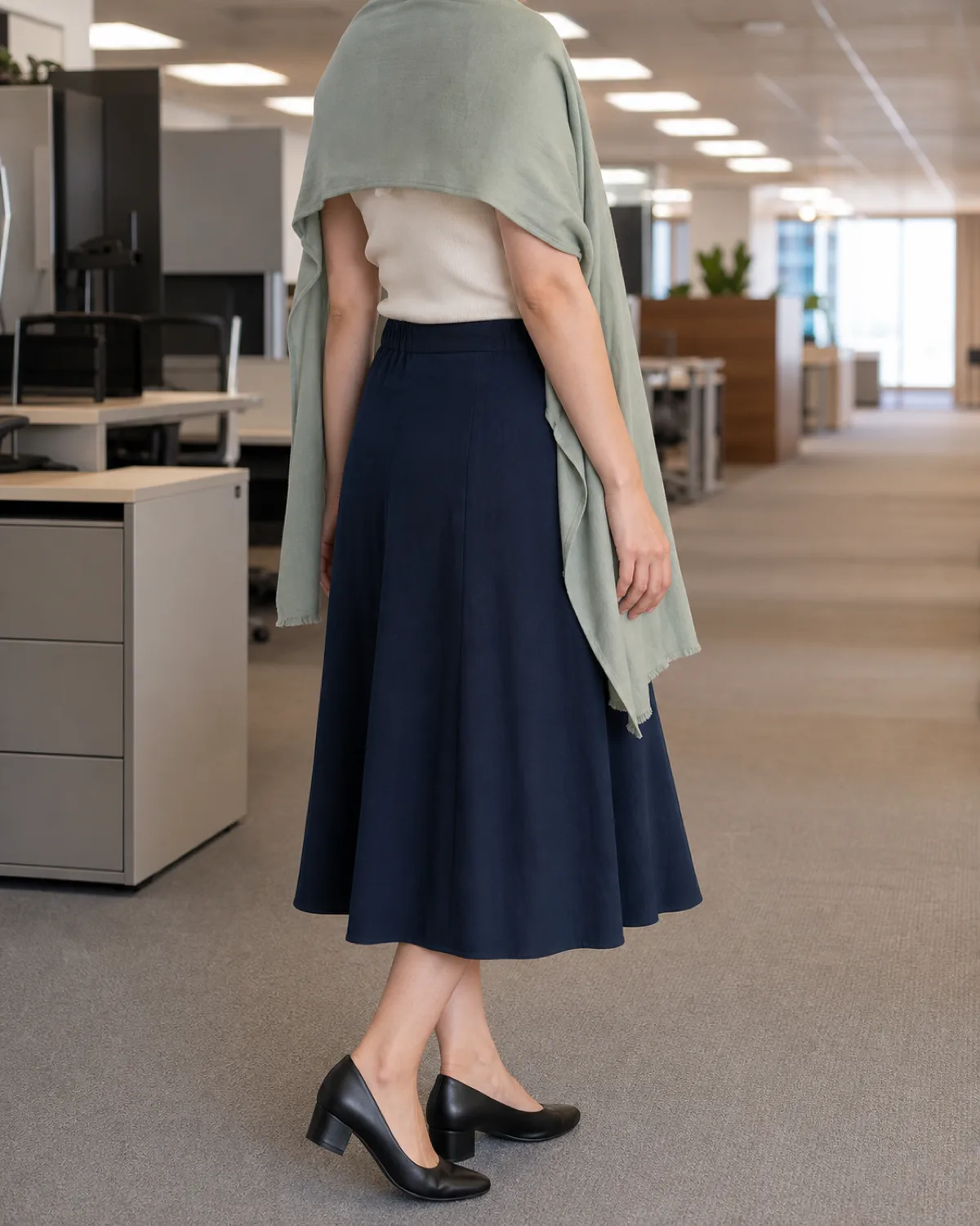A woman seen from behind in a muted sage cotton-wool scarf draped over her shoulders with an ivory knit shell and navy midi skirt in an open-plan office