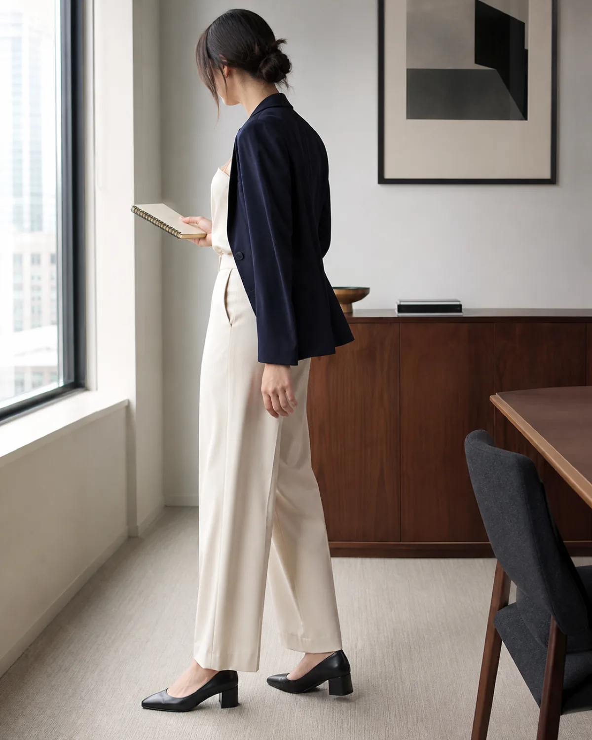 A woman seen from behind in a navy blazer over an ivory silk camisole and cream trousers standing by a window in a modern office