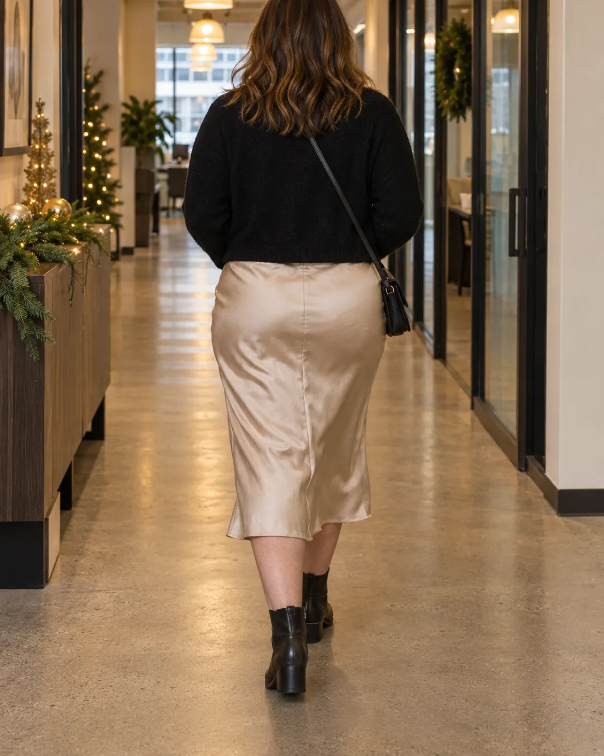 A midsize woman walking away down an office hallway with pendant lights wearing a champagne satin midi slip dress with a cropped black knit cardigan over it and a black crossbody bag with dark leather ankle boots