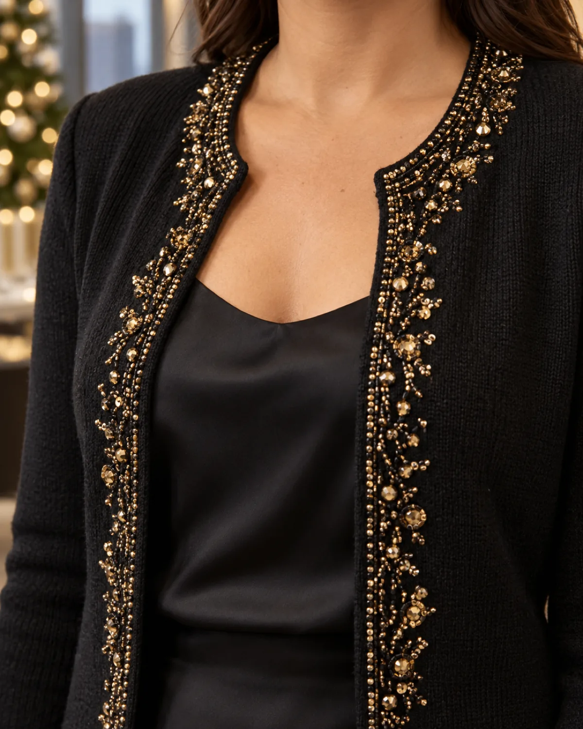Close crop on the shoulder and upper chest of a midsize woman wearing a black cardigan with gold bead and crystal embellishment along the open front panels over a black silk camisole with thin straps visible underneath