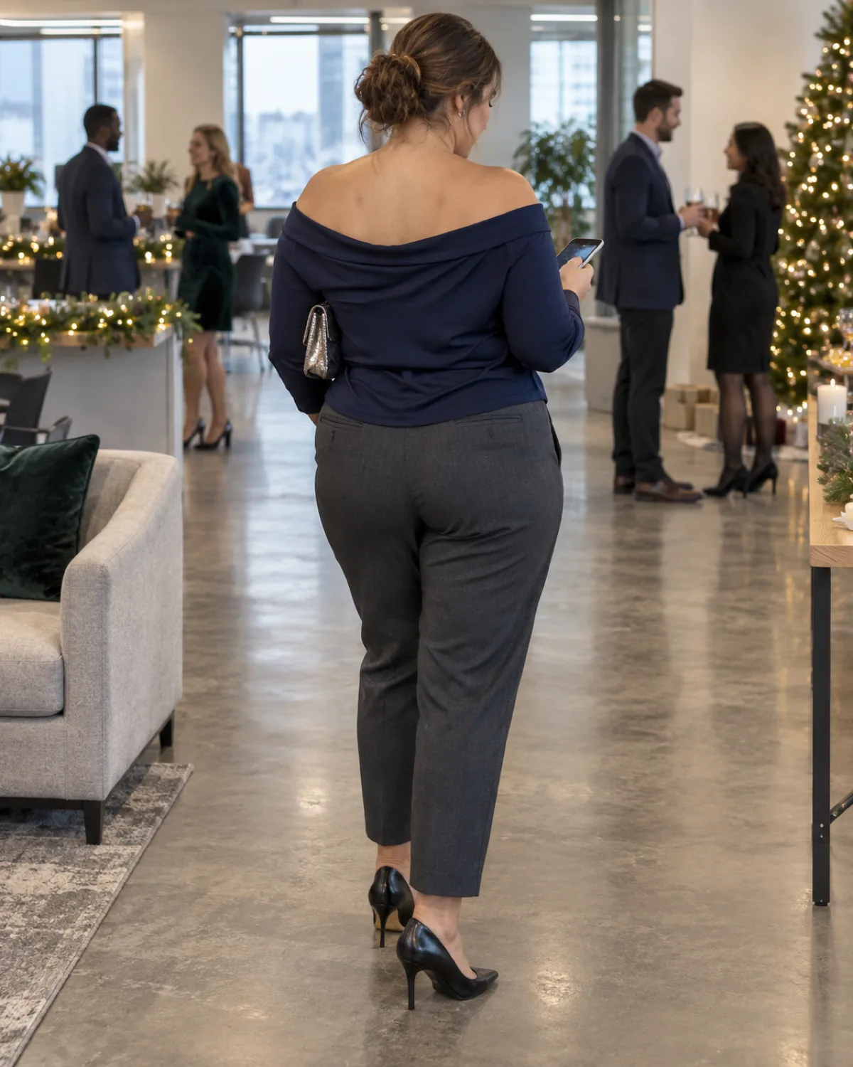 A midsize woman seen from behind at three-quarter angle looking at her phone in an open-plan office with people mingling near a Christmas tree wearing a navy off-shoulder draped top and charcoal tapered ankle trousers with black pumps and a metallic bag tucked under her arm