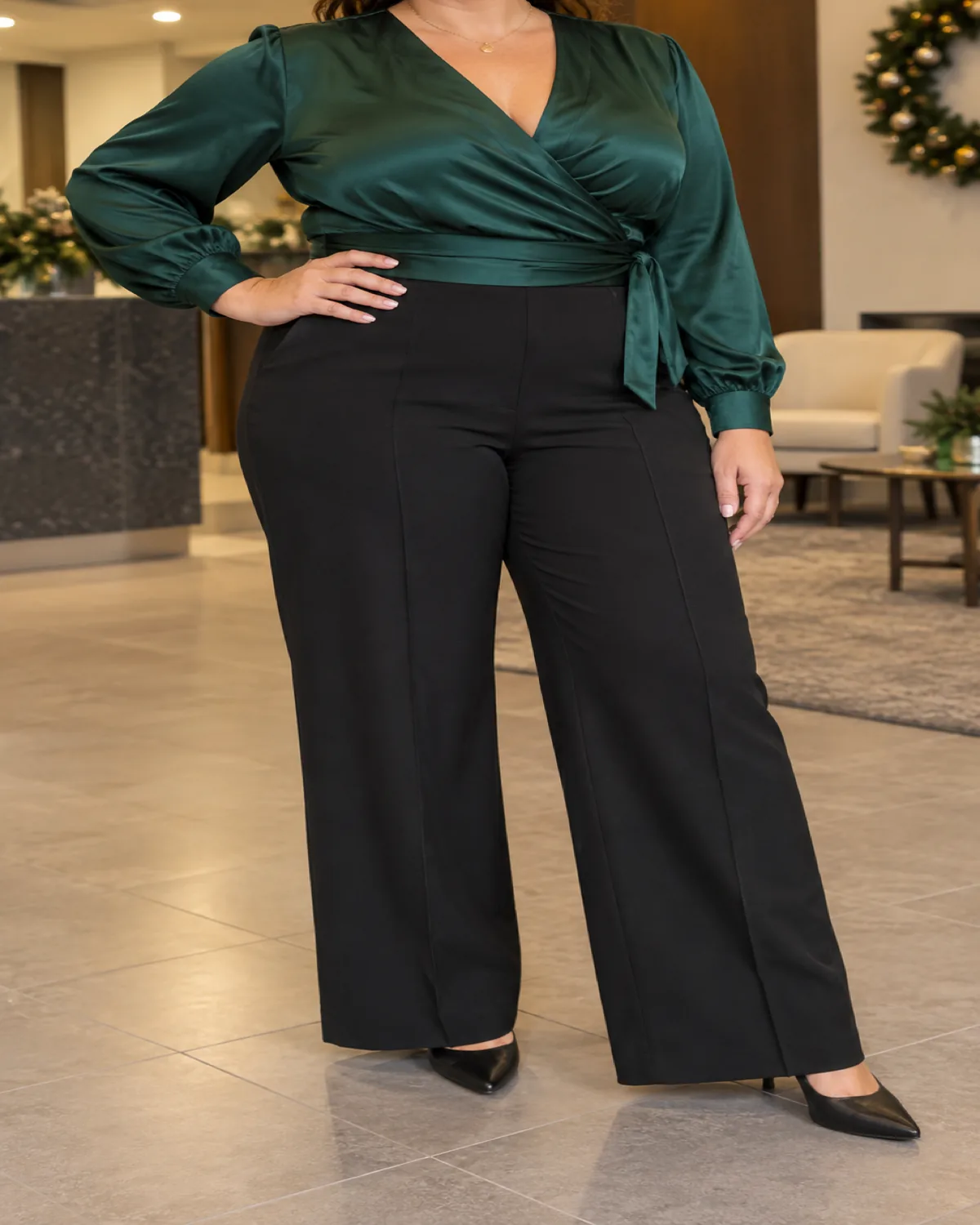 A midsize woman at three-quarter turn in an office lobby with a holiday wreath wearing an emerald satin wrap top tied at the waist and high-waist black wide-leg trousers with black pointed-toe pumps