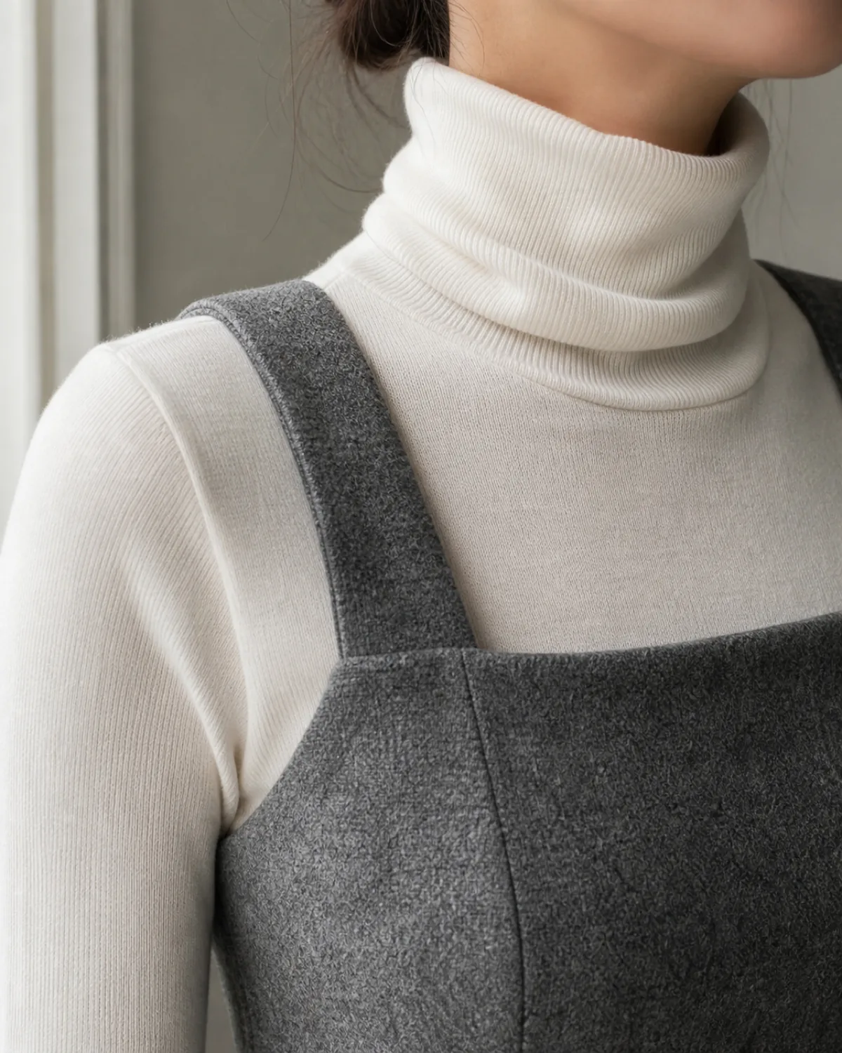 Close crop on the shoulder area of a grey flannel pinafore dress with wide straps over a white fine-knit turtleneck, showing the layered textures