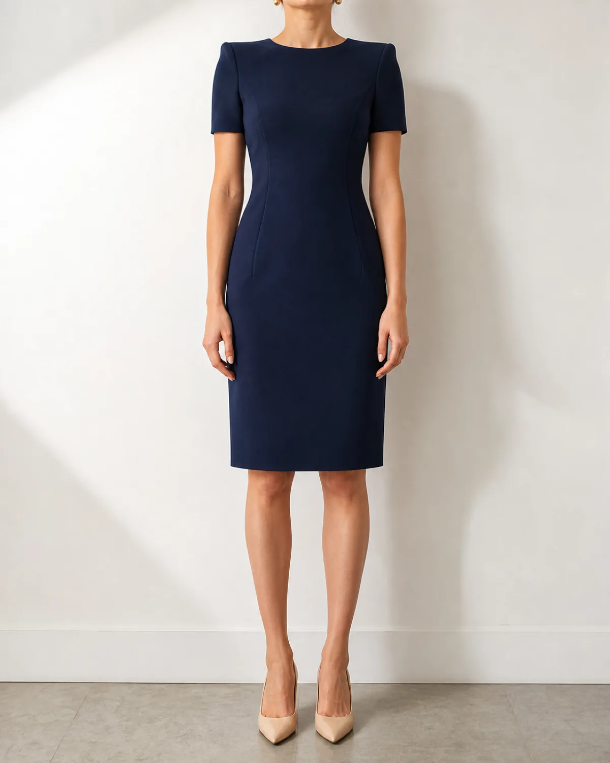 A woman in a navy crepe sheath dress at knee length with nude pointed-toe pumps, standing straight-on against a white wall