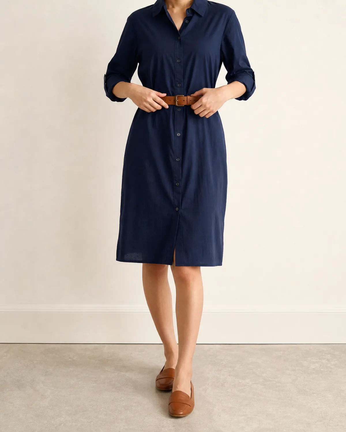 A woman in a navy cotton shirt dress with a collar and button-front placket, a tan leather belt at the waist, hands adjusting the belt, mid-torso crop