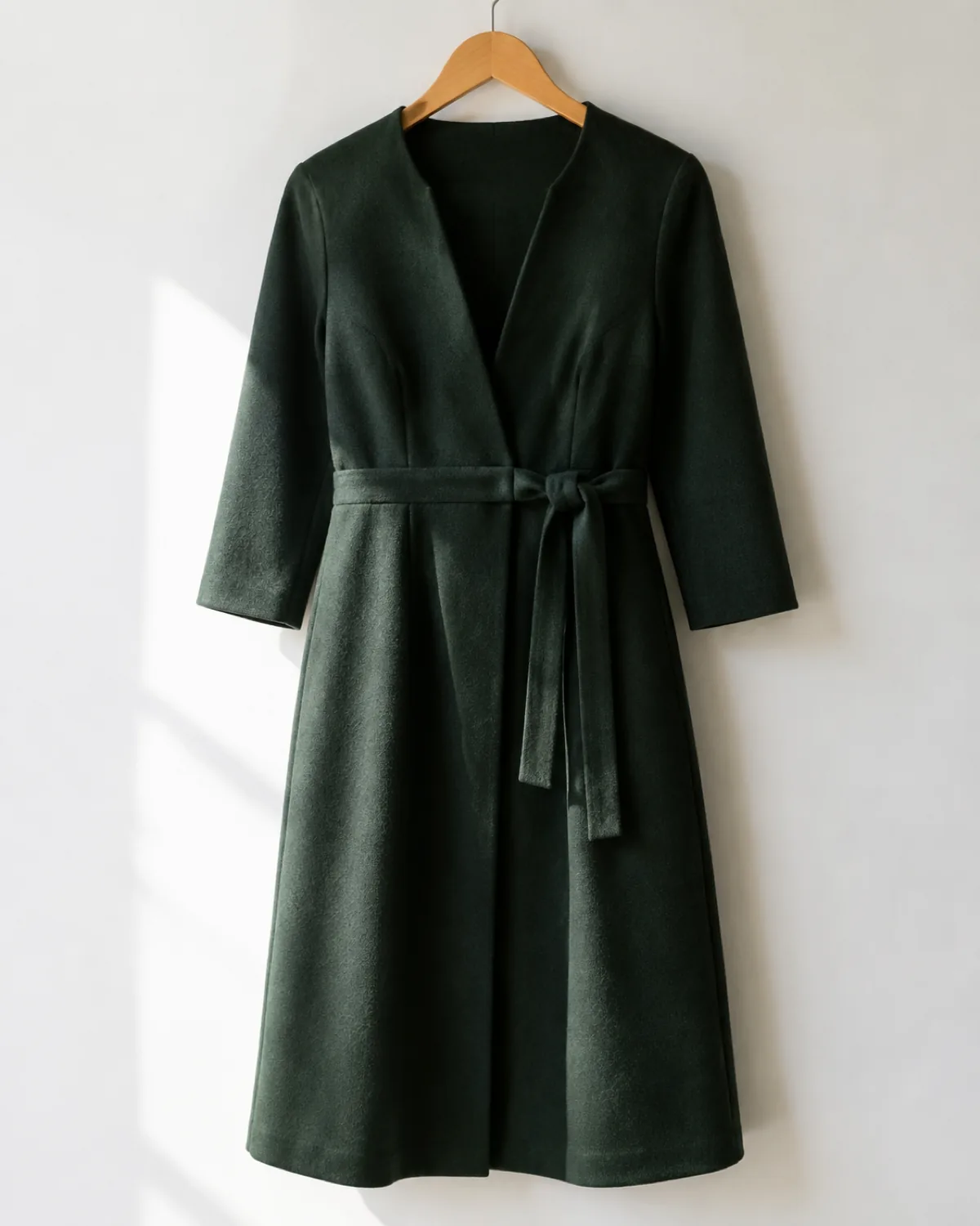 A forest green collarless coat-dress on a wooden hanger against a white wall, showing the self-fabric waist-tie at the empire line, three-quarter sleeves, and V-shaped open neckline