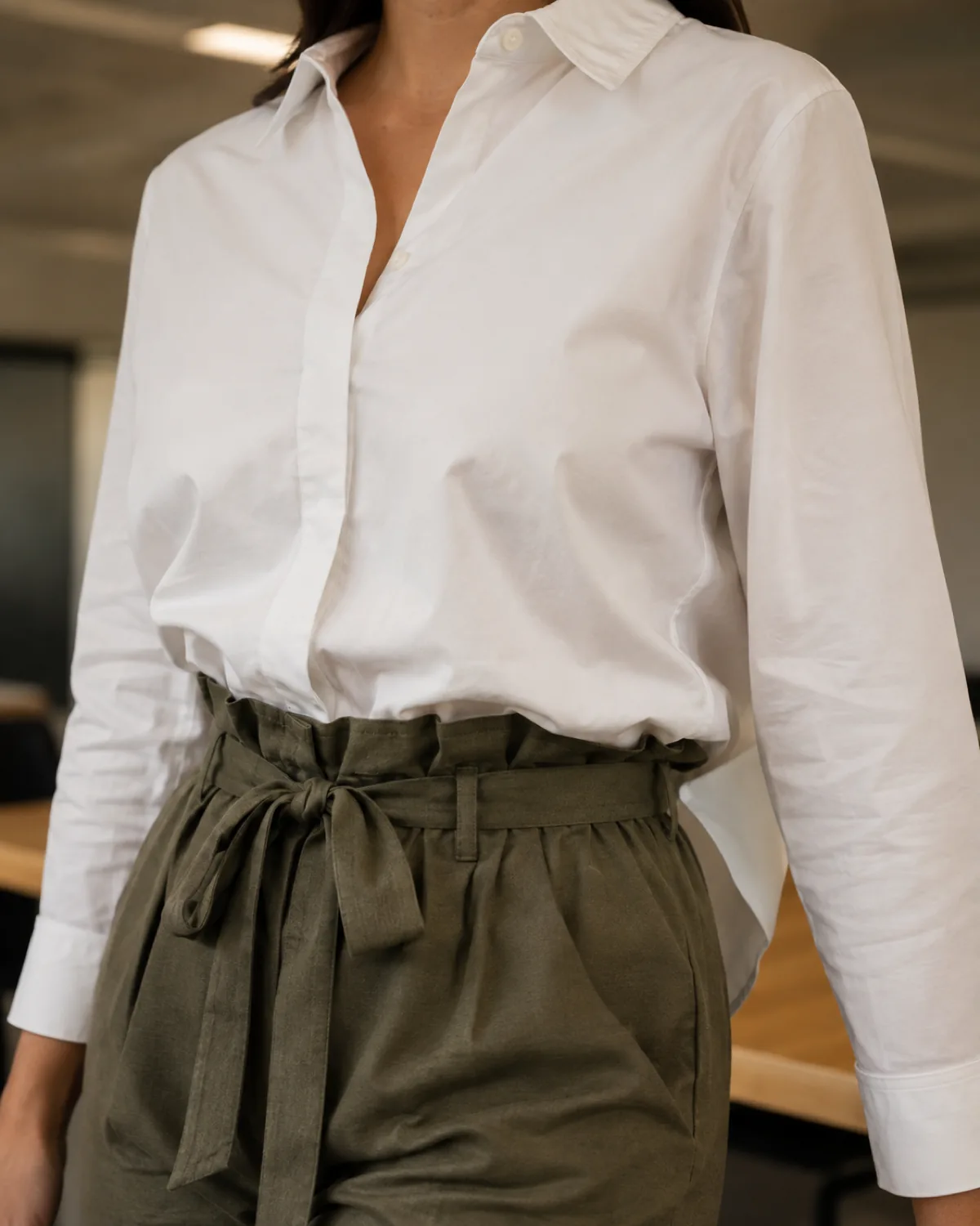 A close-up of a woman's shoulder and collar area showing a white cotton shirt French-tucked into olive green paperbag-waist trousers, with the gathered waistband and fabric tie bow clearly visible