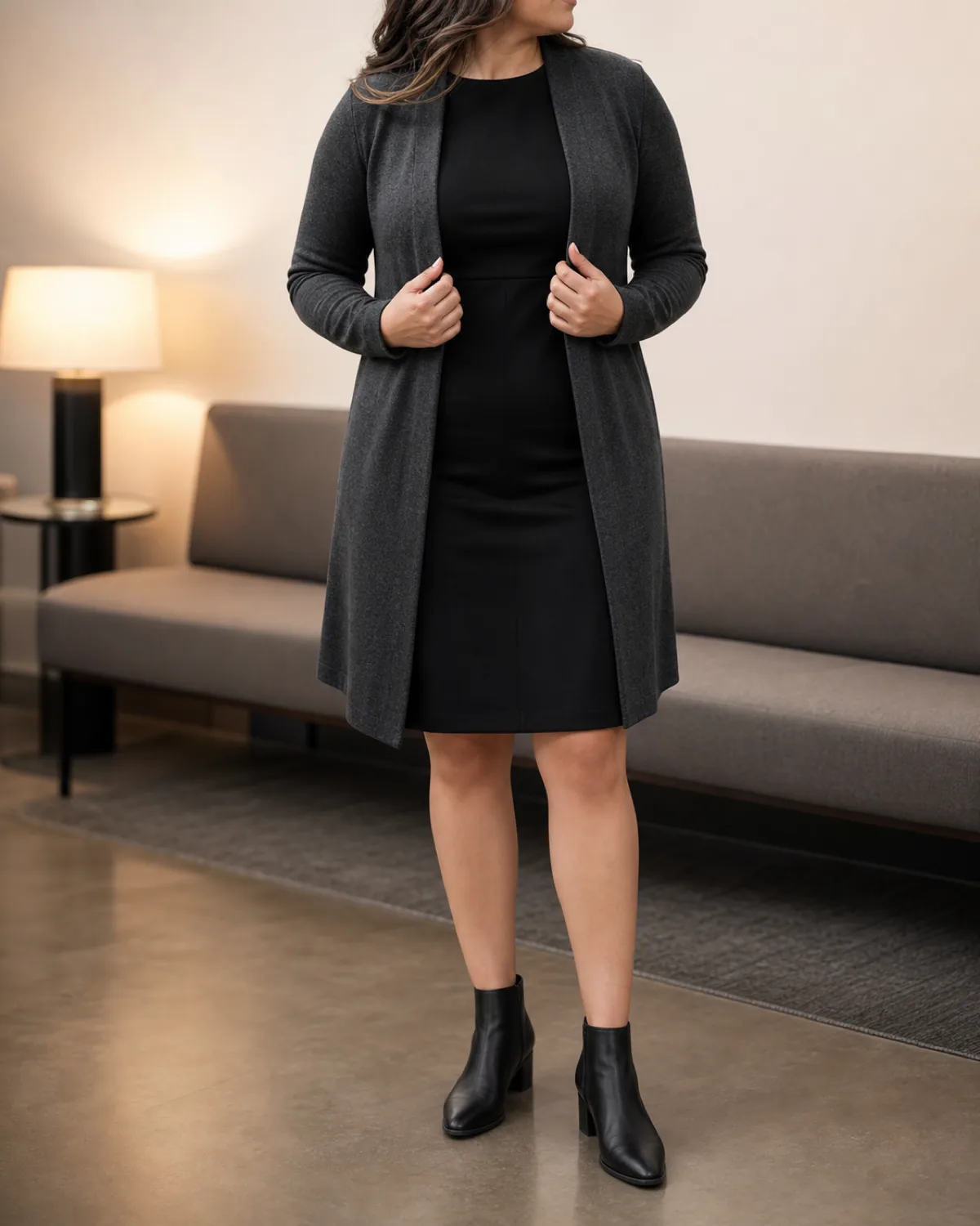 A woman with an apple body shape in a mid-torso crop adjusting the open front of a charcoal longline cardigan over a black ponte sheath dress, with black leather ankle boots, in a minimalist office lounge