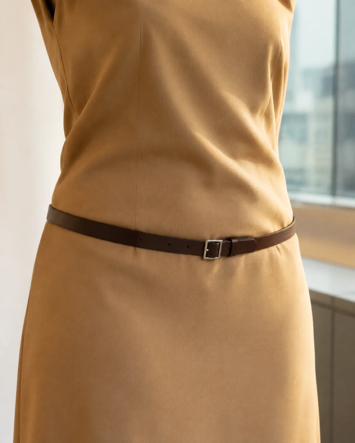 A close-up of a woman's waist wearing a camel-coloured shift dress with a narrow dark brown leather belt placed at the high hip, showing the small plain square buckle and smooth fabric drape