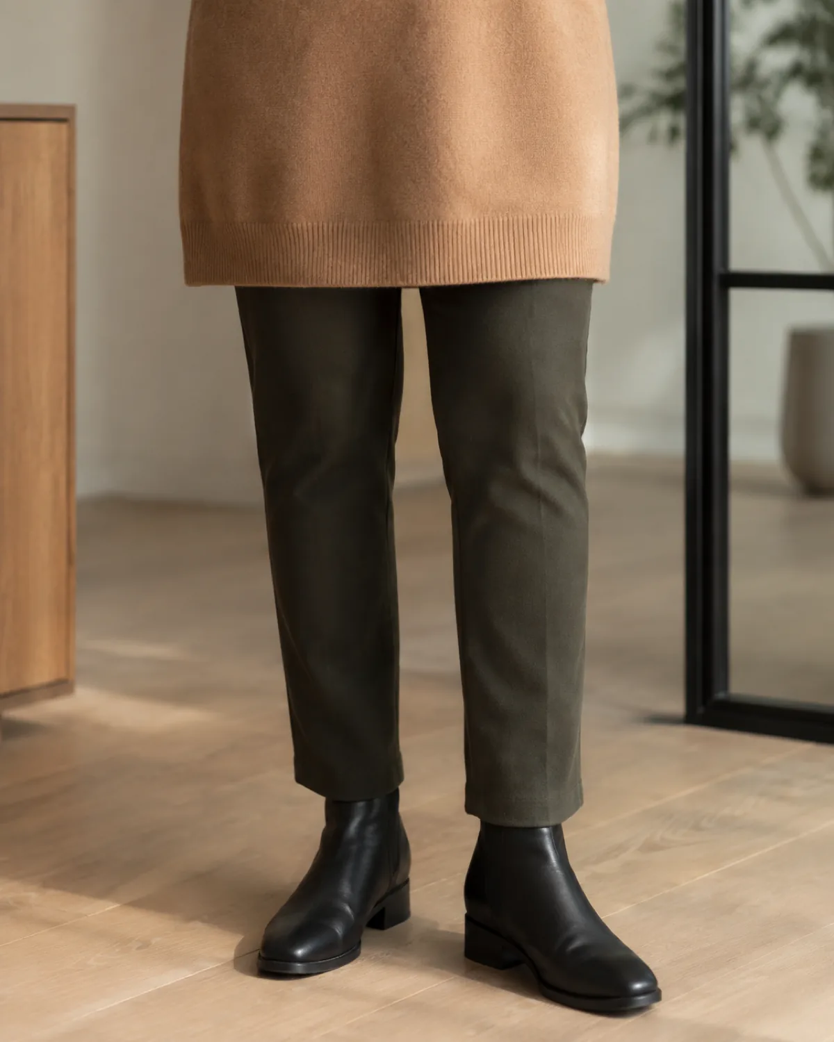 A close-up of the lower half of an outfit showing a camel cashmere tunic sweater hem ending at mid-thigh, dark olive slim-cut trousers, and black leather ankle boots on a light wood office floor