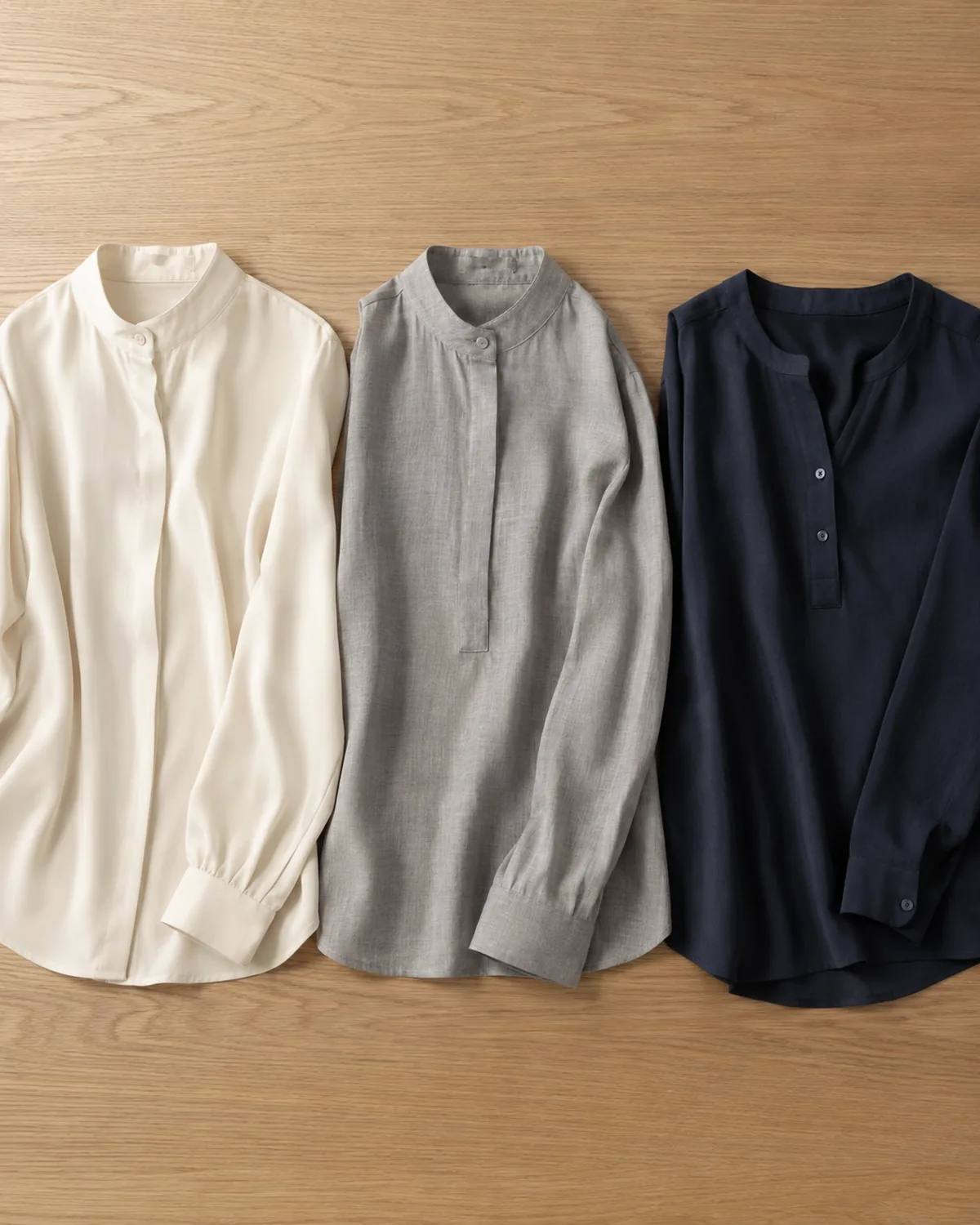 Three professional blouses in flat-lay on a light oak surface, a cream mandarin-collar blouse on the left, a light grey band-collar blouse in the centre and a navy popover blouse on the right