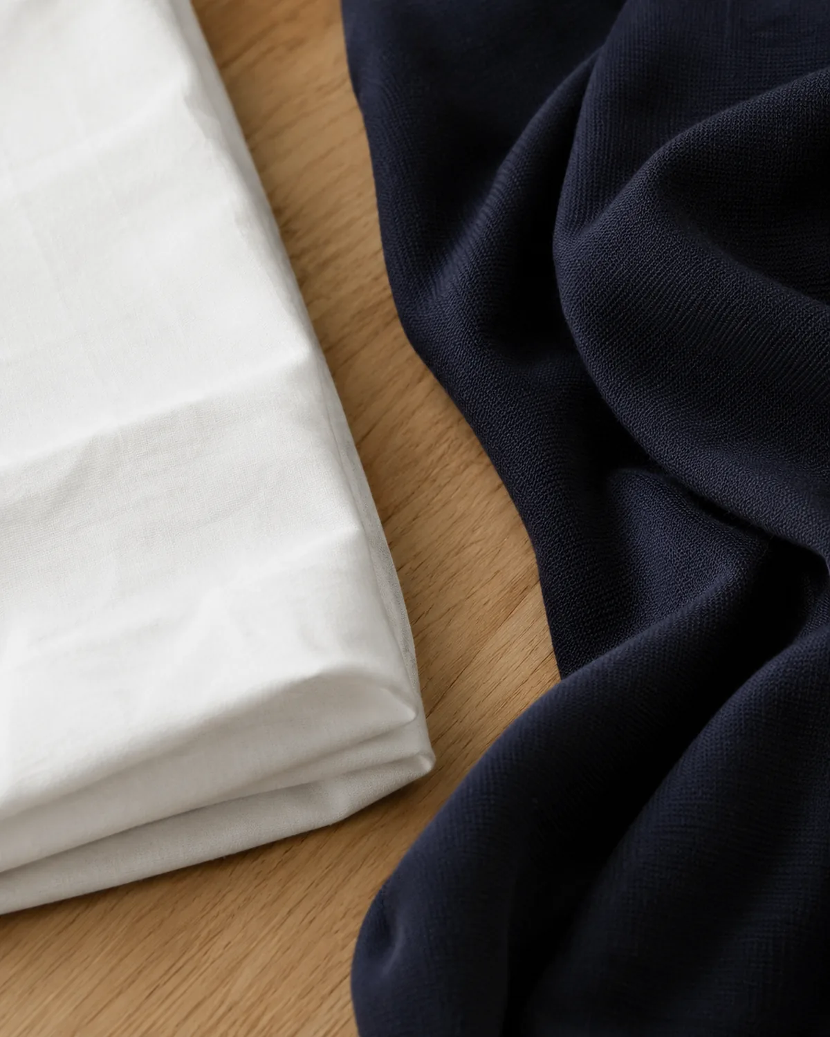 Two pieces of fabric on a light oak surface, white poplin folded flat and rigid on the left, navy ponte knit draped softly showing natural folds on the right