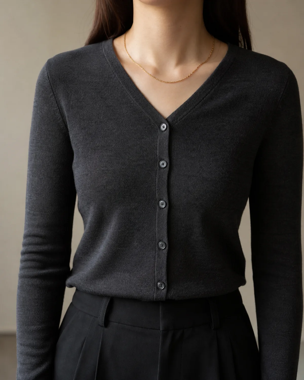 Close-up of a charcoal V-neck fine-knit cardigan buttoned fully closed as a top layer over black trousers, with a thin gold chain necklace