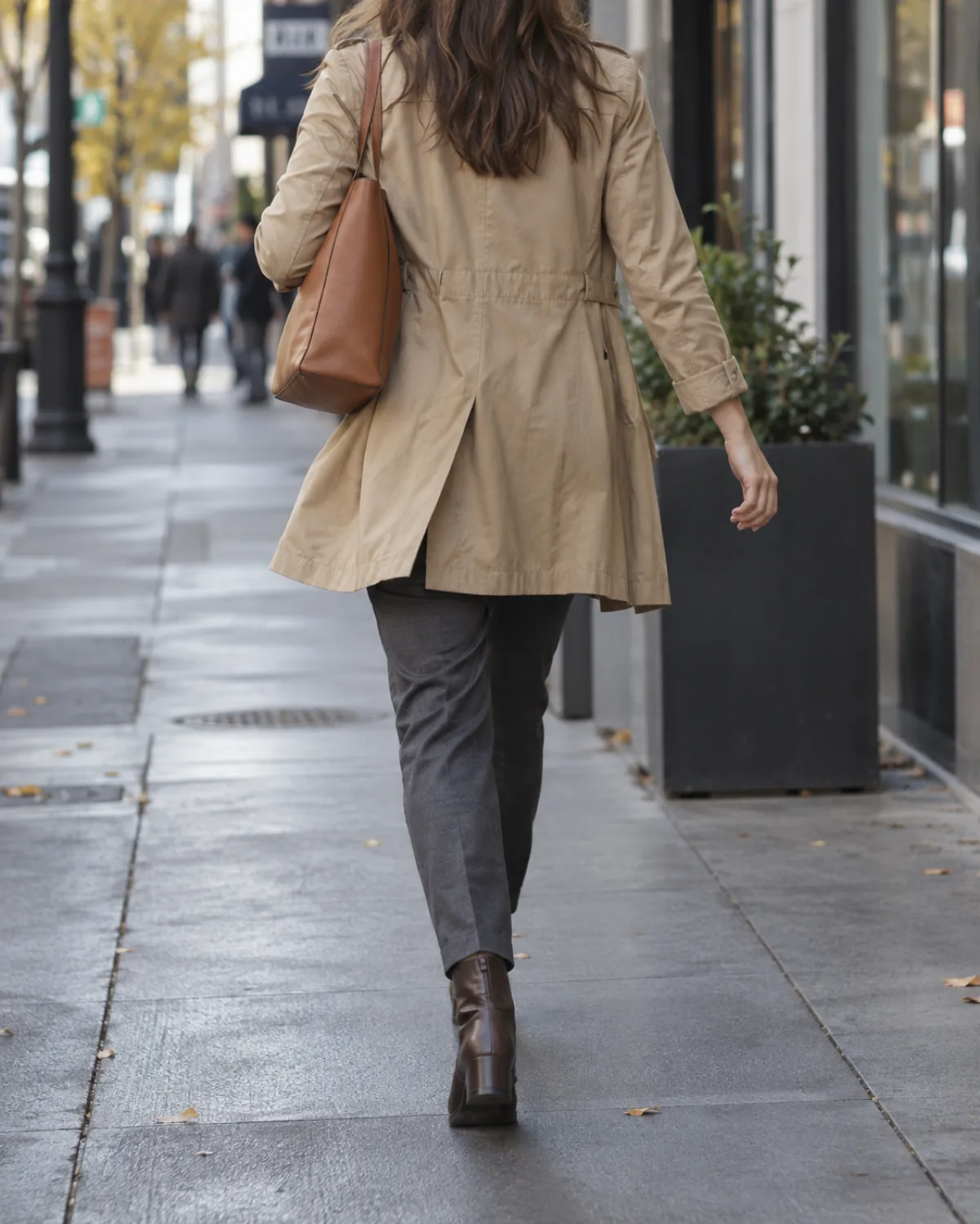A woman walking away from the camera on a city sidewalk in overcast early-morning light wearing a khaki lightweight cotton trench coat worn open over charcoal tapered trousers and dark brown ankle boots with a tan structured leather shoulder bag