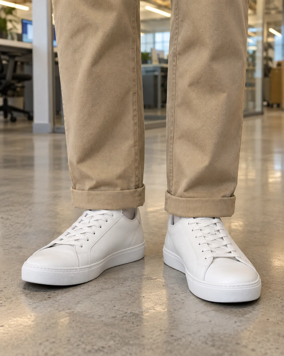 Close-up of the lower legs showing sand-coloured straight-leg khakis cuffed once above clean white leather low-top sneakers on a polished concrete office floor