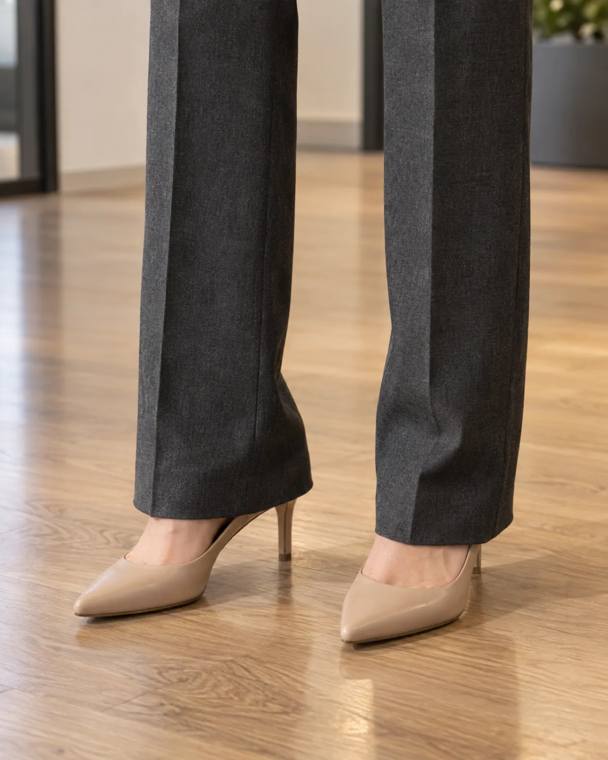 Close crop from mid-calf down showing charcoal wool trousers with a clean front crease breaking slightly over nude pointed-toe pumps with a mid-height stiletto heel on a polished light wood office floor