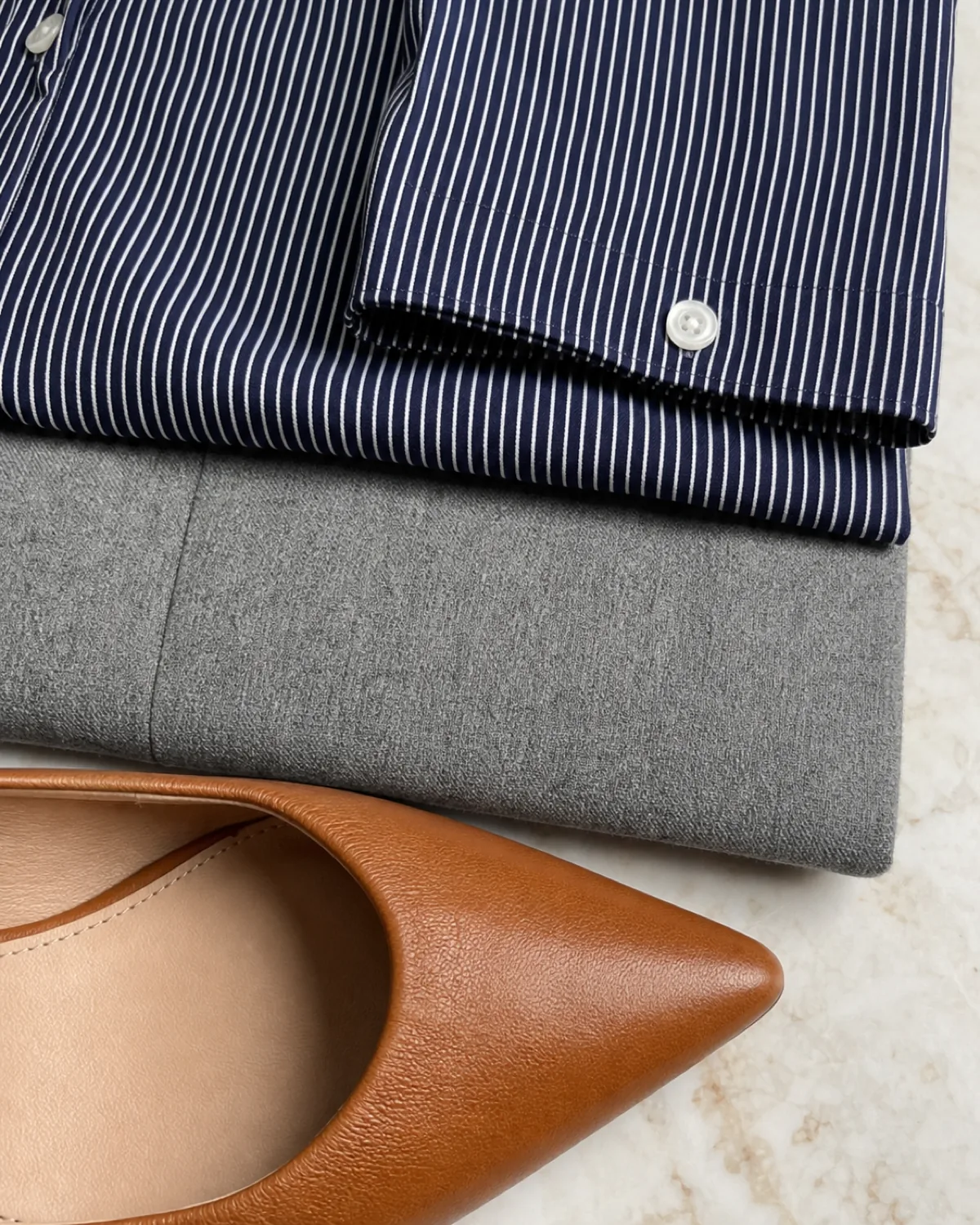 Stacked macro detail of a folded navy-and-white pinstripe cotton button-down shirt on top, a folded light grey wool trouser fabric in the middle, and the toe of a cognac leather pointed-toe flat at the bottom, on a pale marble surface