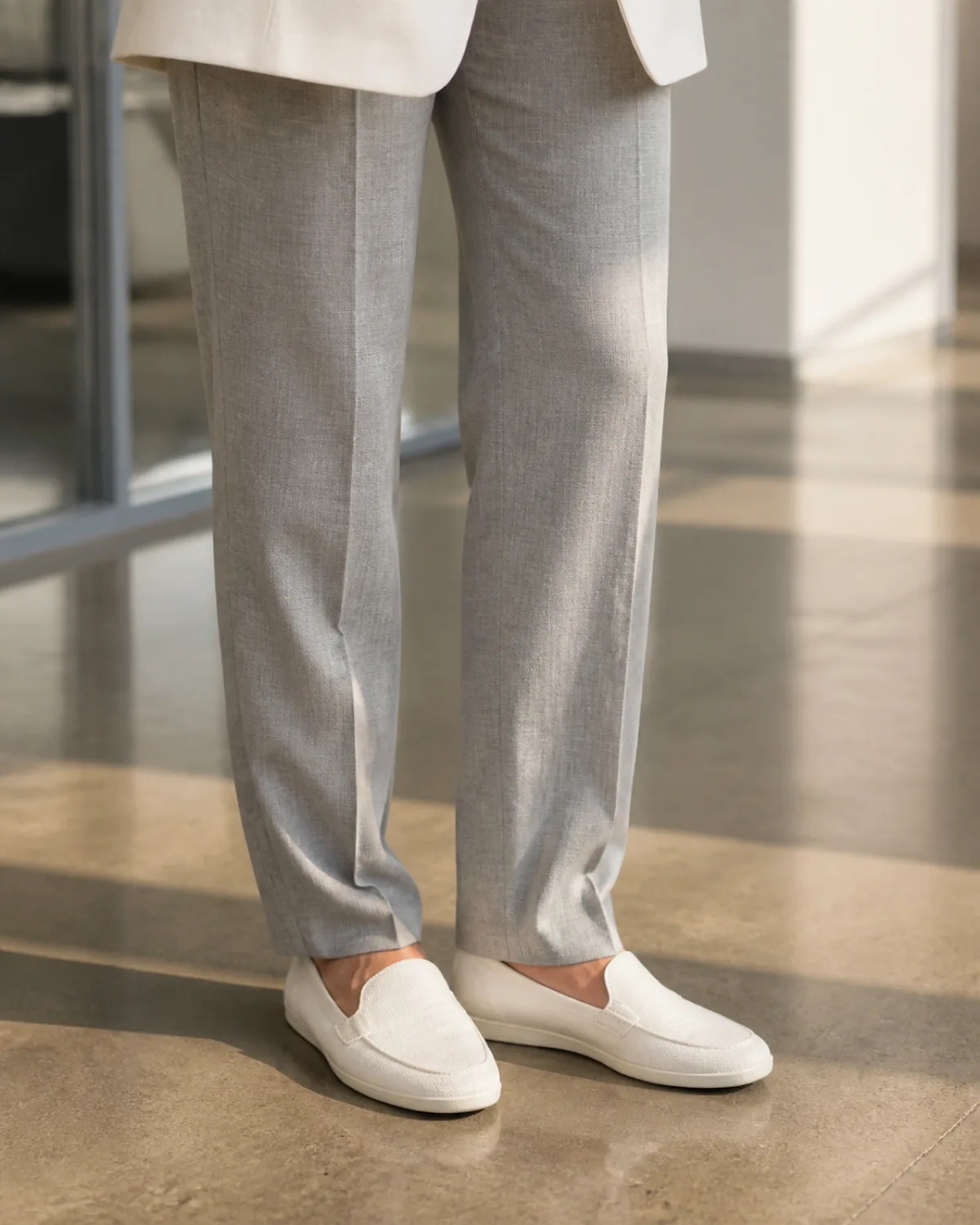 Close crop on the hem and shoes: medium grey tapered linen-blend trousers with a slight break at the ankle, the bottom edge of a white linen blazer visible at top of frame, and white canvas slip-on loafers on a polished concrete floor