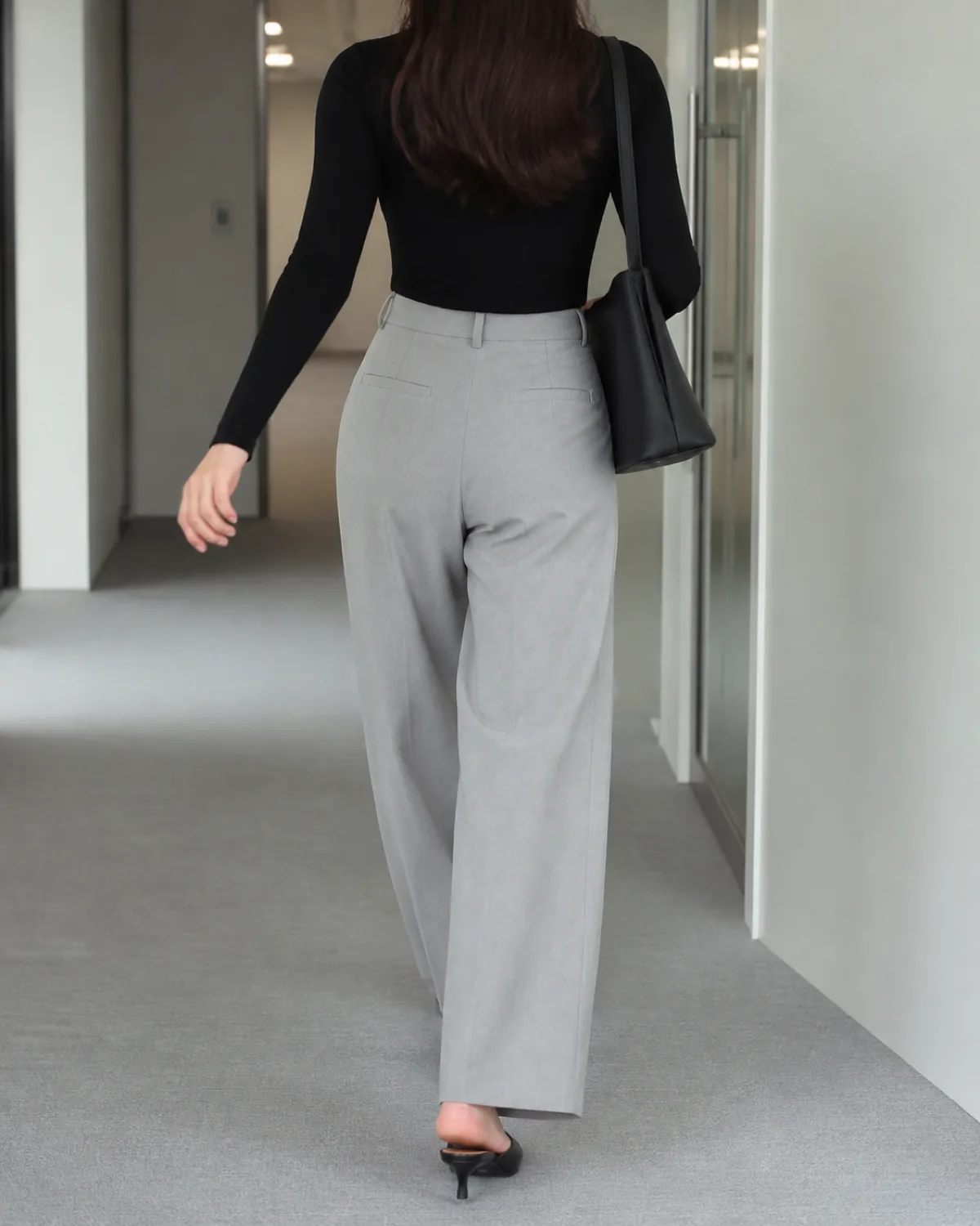 A woman walking away in an office hallway wearing light grey wide-leg trousers, a fitted black turtleneck tucked in, a black structured shoulder bag, and black pointed mules