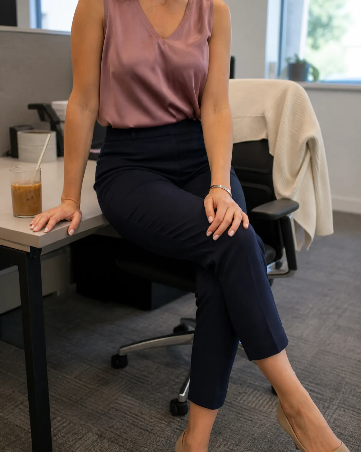 A woman seated at the edge of a desk in a dusty-rose sleeveless shell tucked into navy ankle trousers with a cream lightweight knit cardigan draped over the desk chair behind her and an iced coffee on the desk