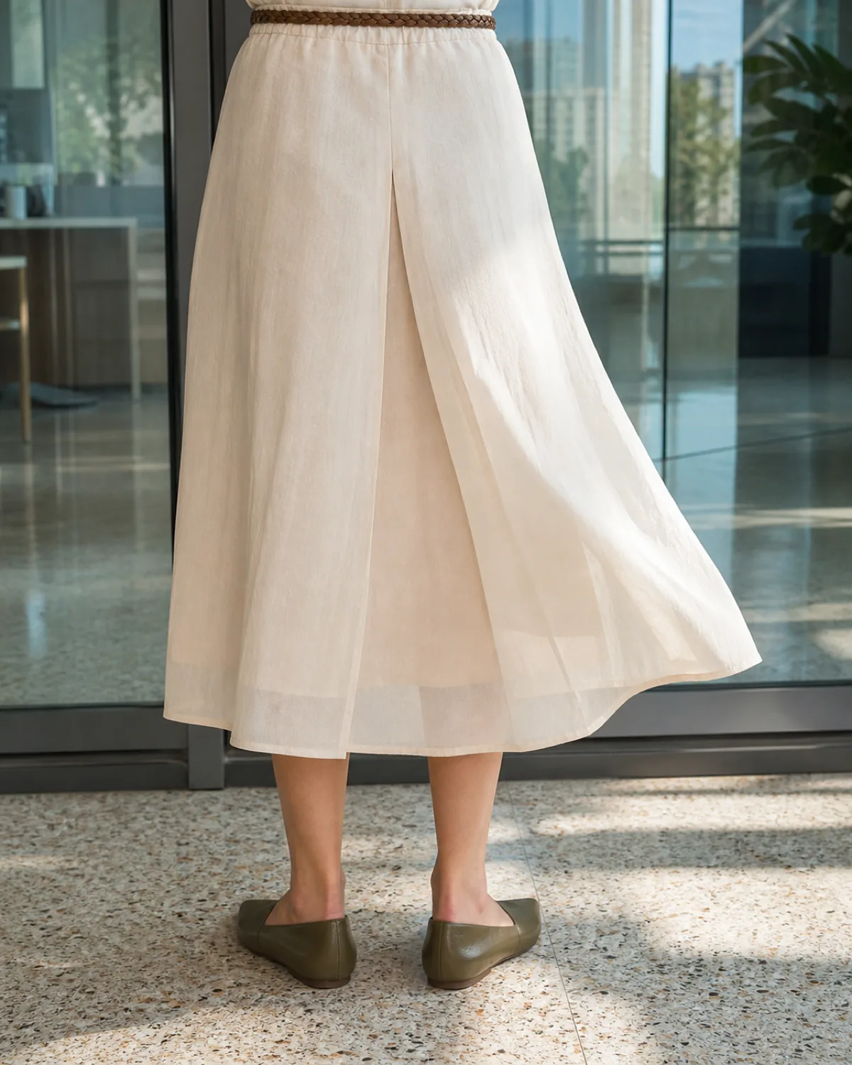 A close crop on the lower body showing a cream cotton-voile midi skirt with a visible back pleat catching light, a braided leather belt at the waist, bare ankles, and olive-green pointed-toe leather flats on a terrazzo office floor with a glass door reflecting daylight
