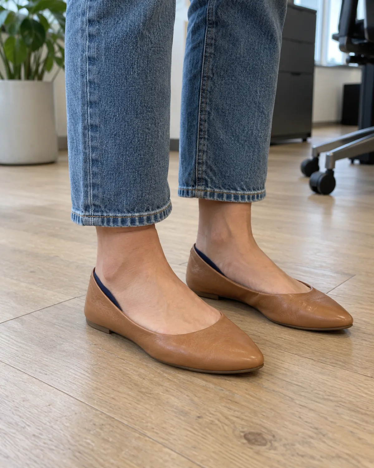 A close crop from mid-calf down showing cropped straight-leg medium-wash jeans hemmed two inches above the ankle with tan leather ballet flats on a light wood office floor