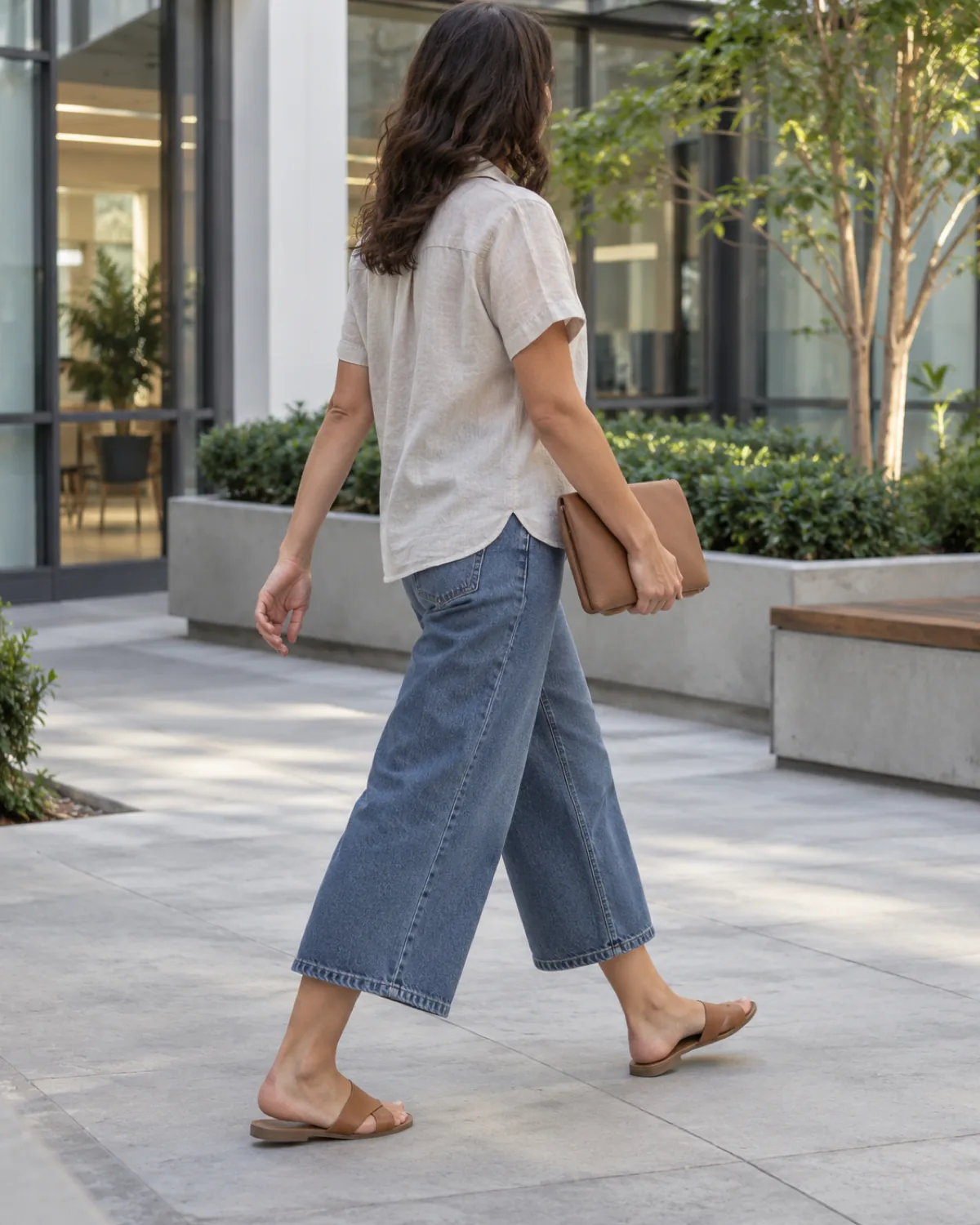 A woman walking away through an outdoor office courtyard with clean architectural lines and greenery wearing cropped wide-leg medium-wash jeans with an untucked oatmeal linen-blend camp shirt and tan leather slide sandals