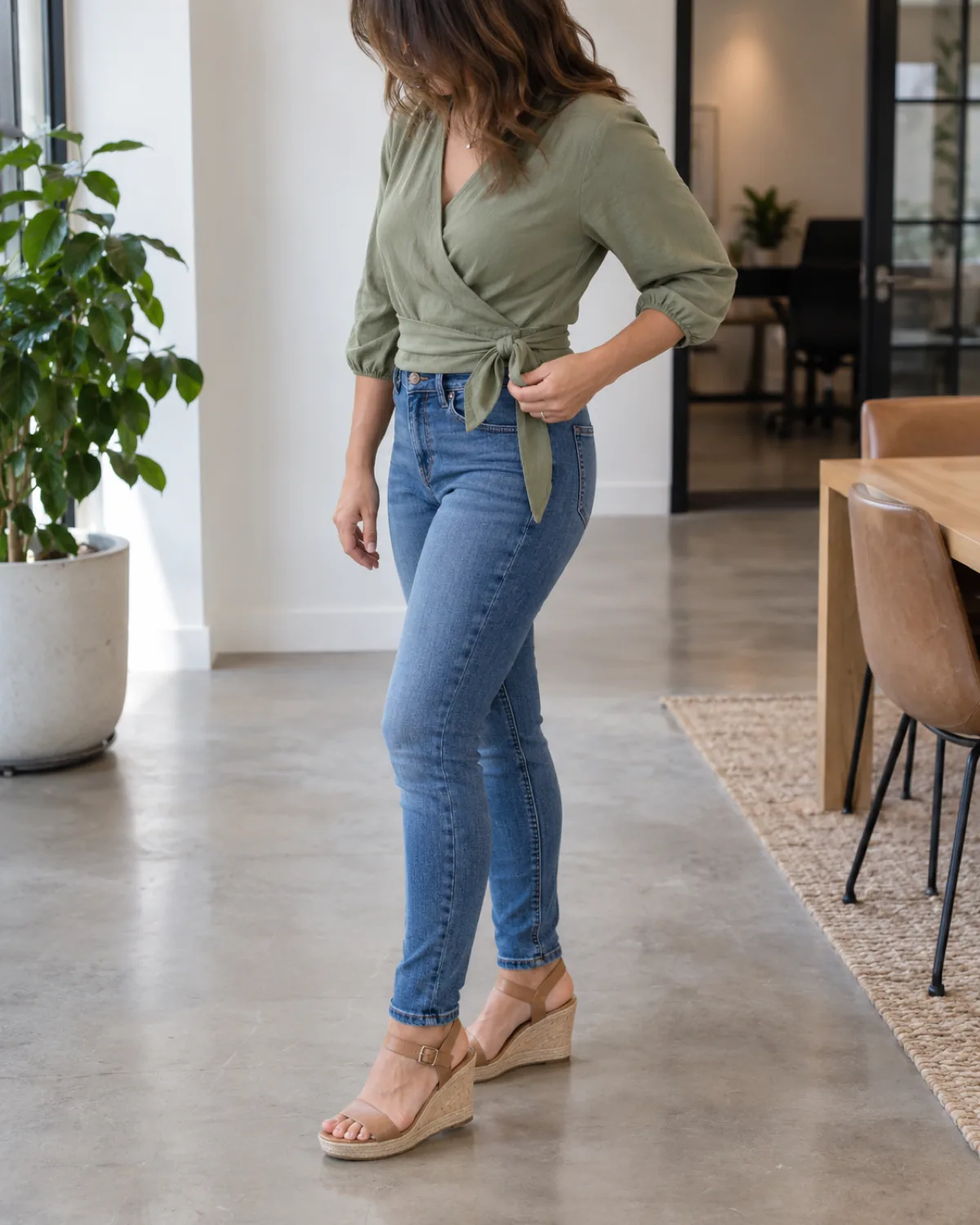A woman in a three-quarter turn to the left wearing slim medium-wash jeans and a sage-green wrap blouse with a side tie tucked into the jeans with tan wedge sandals, adjusting the wrap tie at her hip in a bright office break room