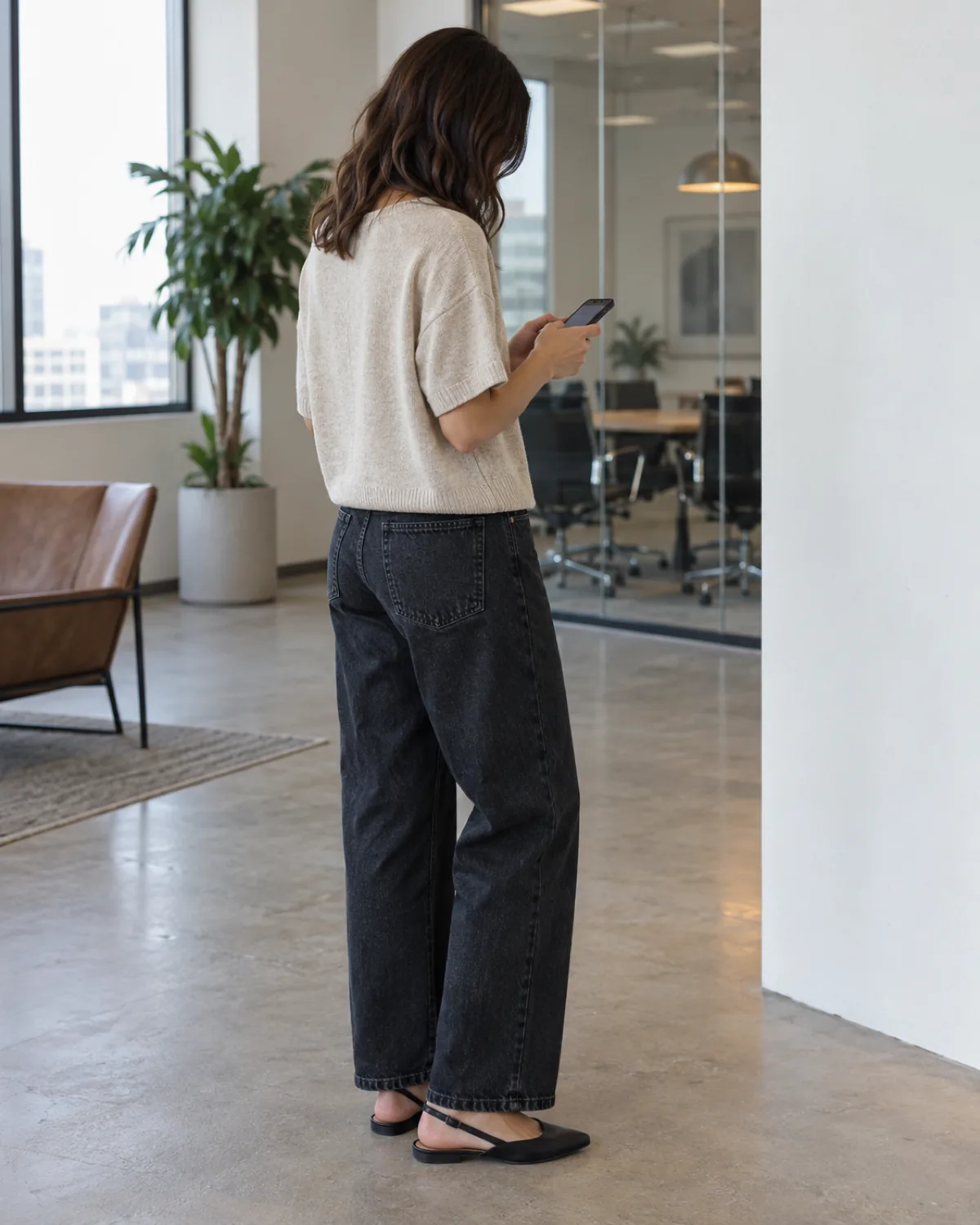A woman seen from a back-three-quarter angle on a rooftop terrace looking down at a phone held at waist height wearing relaxed-fit dark jeans and a boxy oatmeal short-sleeve knit with black slingback flats