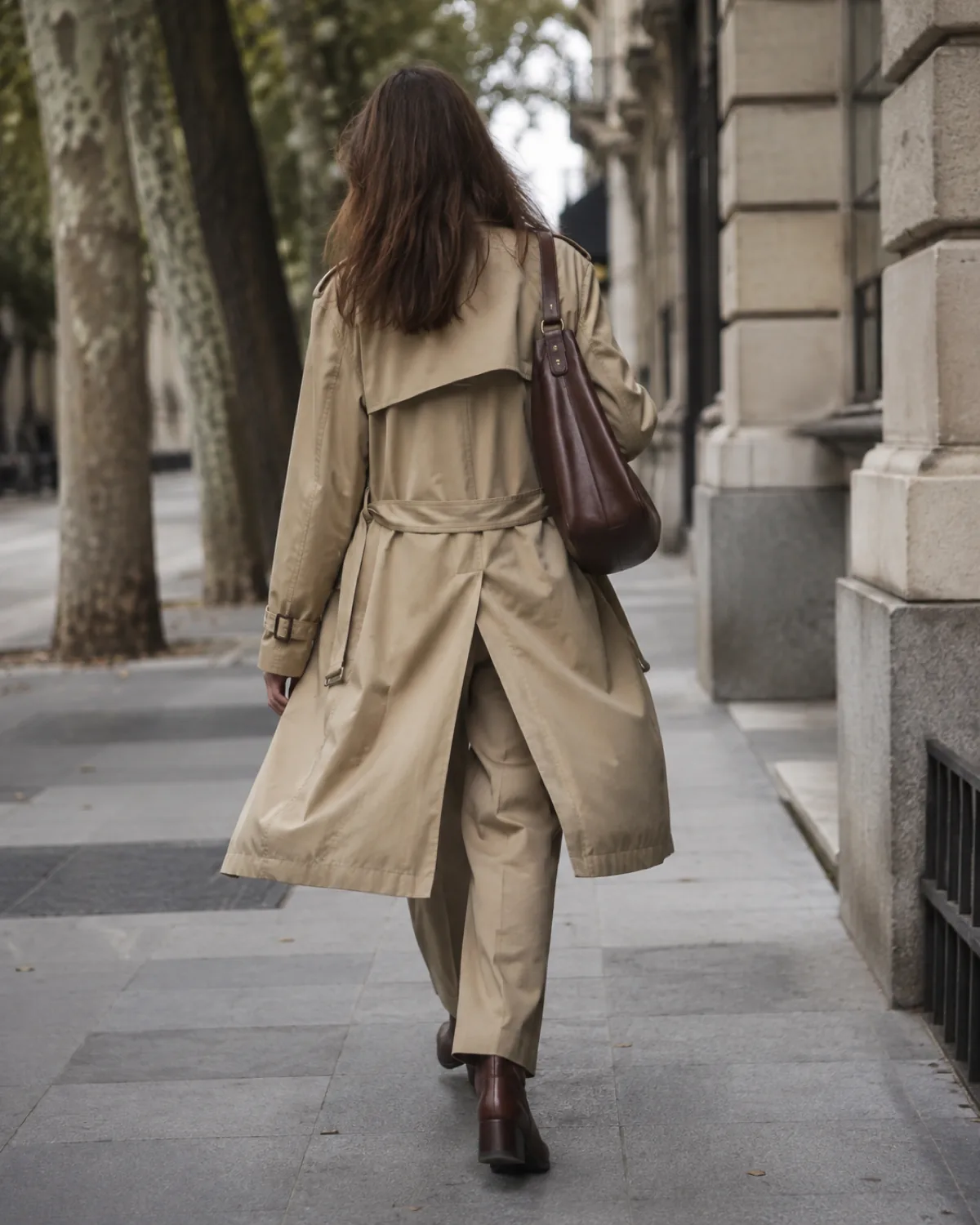 A woman walking away through a city street with trees wearing straight-leg khaki pants, a belted khaki trench coat swinging open, dark brown leather ankle boots, and a structured leather bag on one shoulder