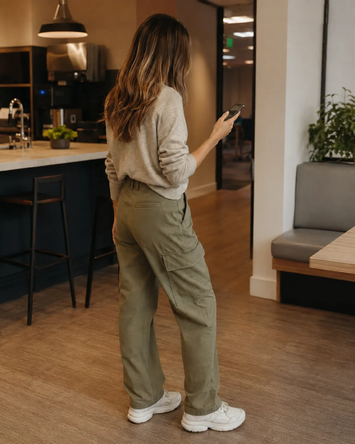 A woman shot from behind in a casual office break room wearing khaki cargo pants with flap side pockets, an oatmeal crewneck sweater tucked in, and chunky off-white sneakers, holding a phone