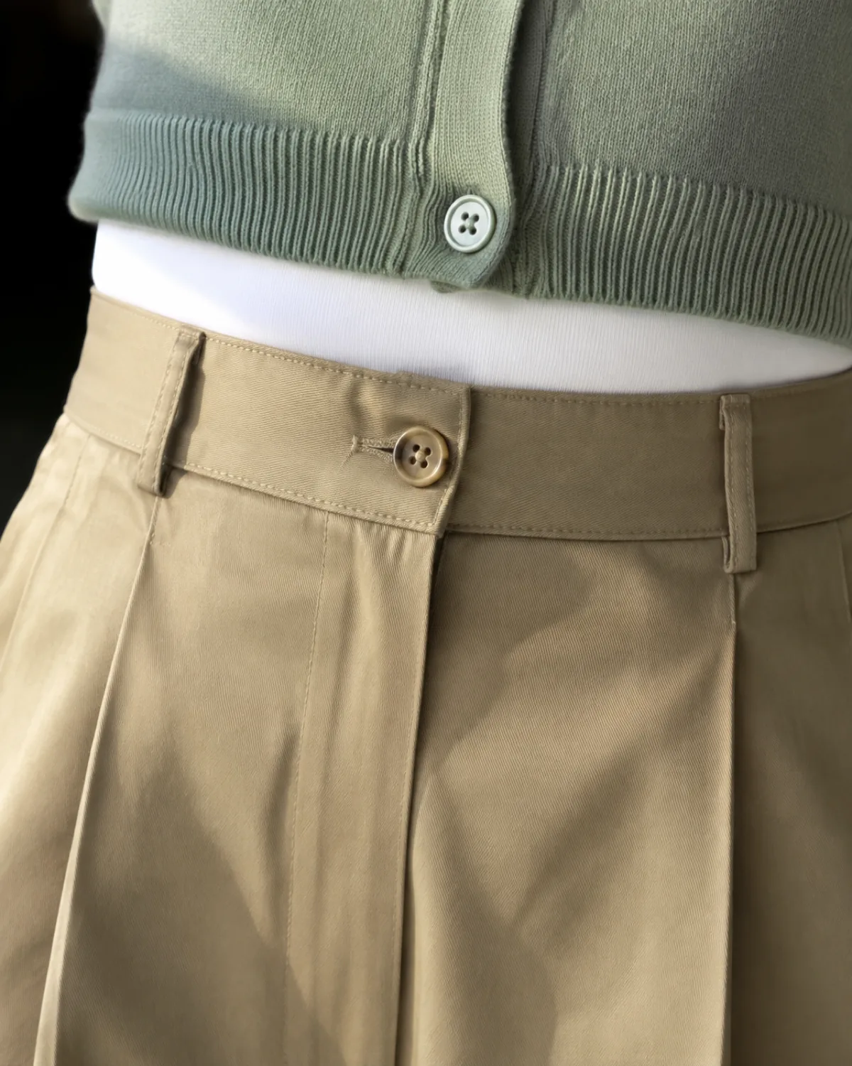 Close-up of a waistline showing wide-leg khaki pants with a high waistband and front pleat, a sage green cropped cardigan ending at the waist, and the tucked edge of a white tank top visible underneath