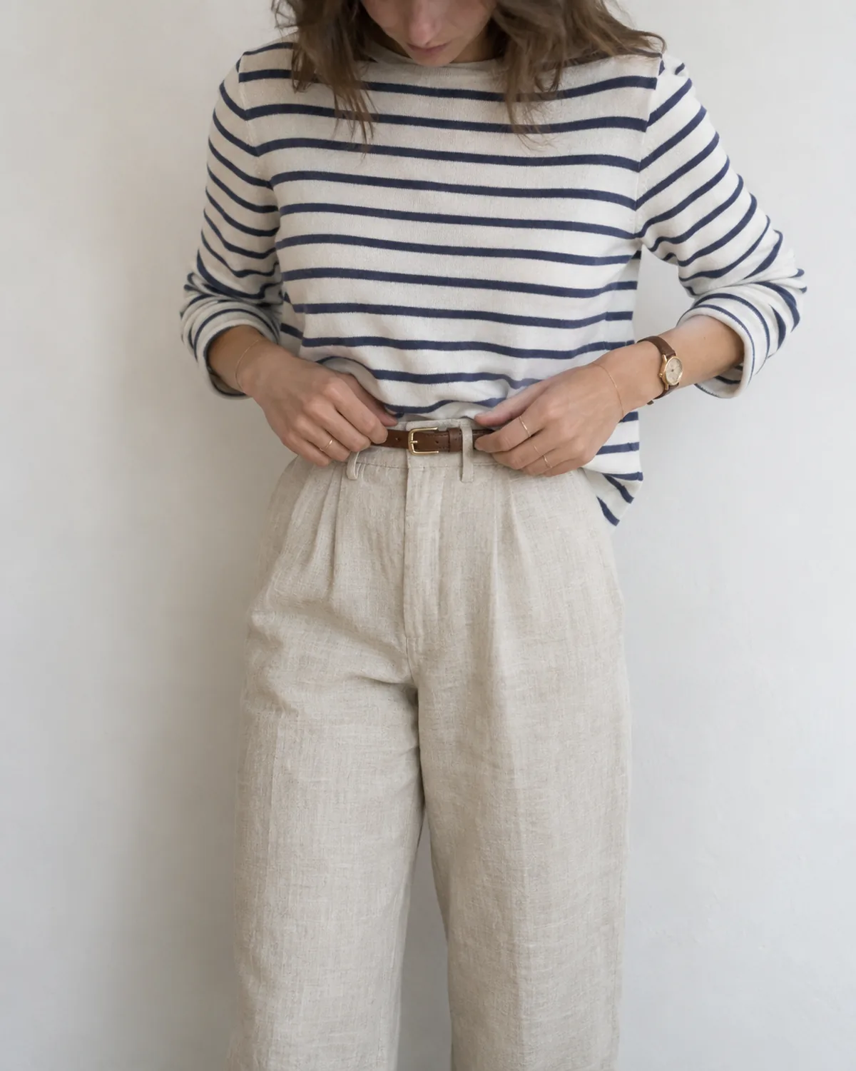 A mid-torso crop showing a white and navy breton-stripe knit half-tucked into high-waisted natural linen trousers, with both hands adjusting a thin brown leather belt