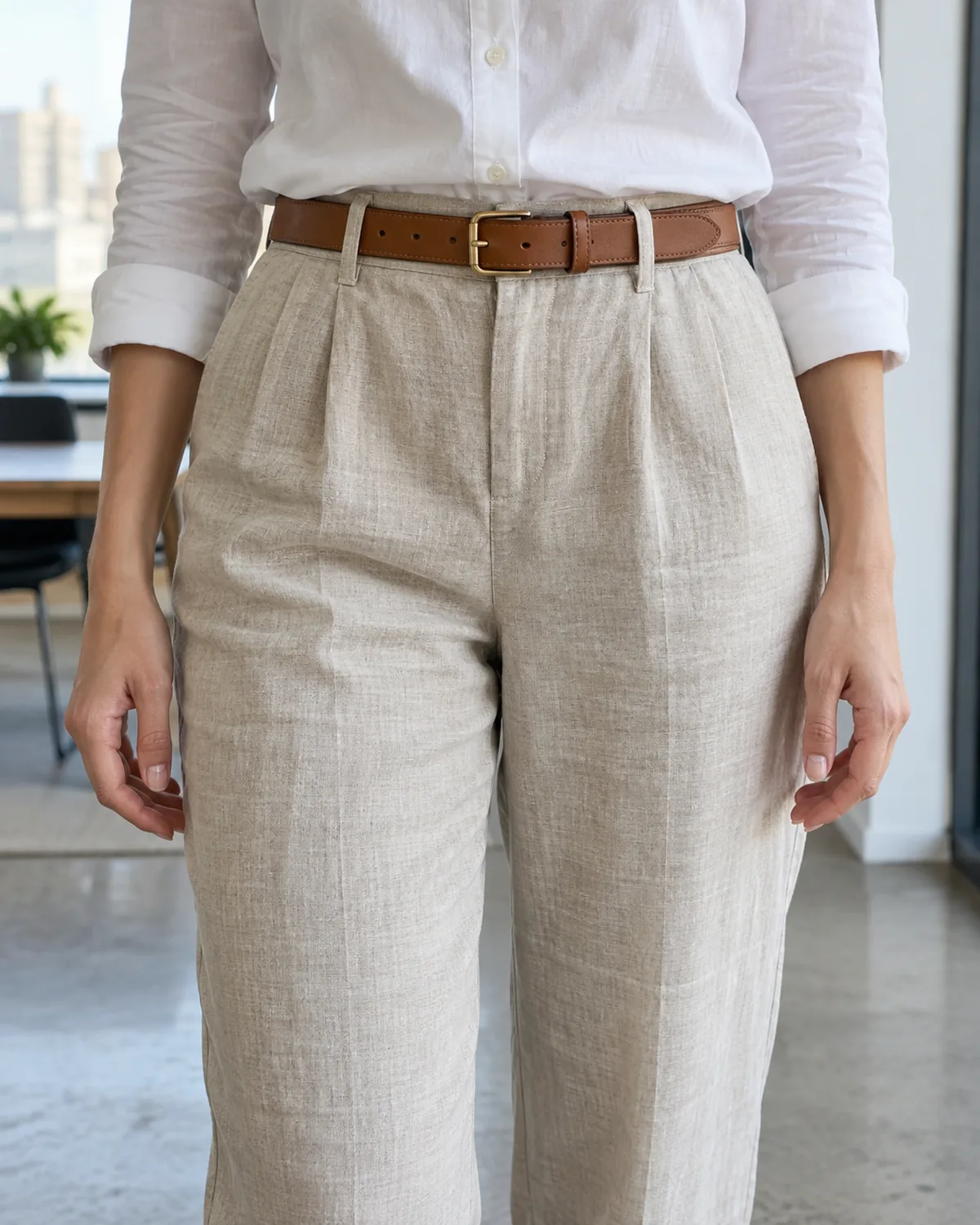 A close crop of the waist area showing natural linen drawstring trousers with a white cotton shirt tucked in and a tan leather belt worn over the shirt at the waistband, the drawstring completely hidden