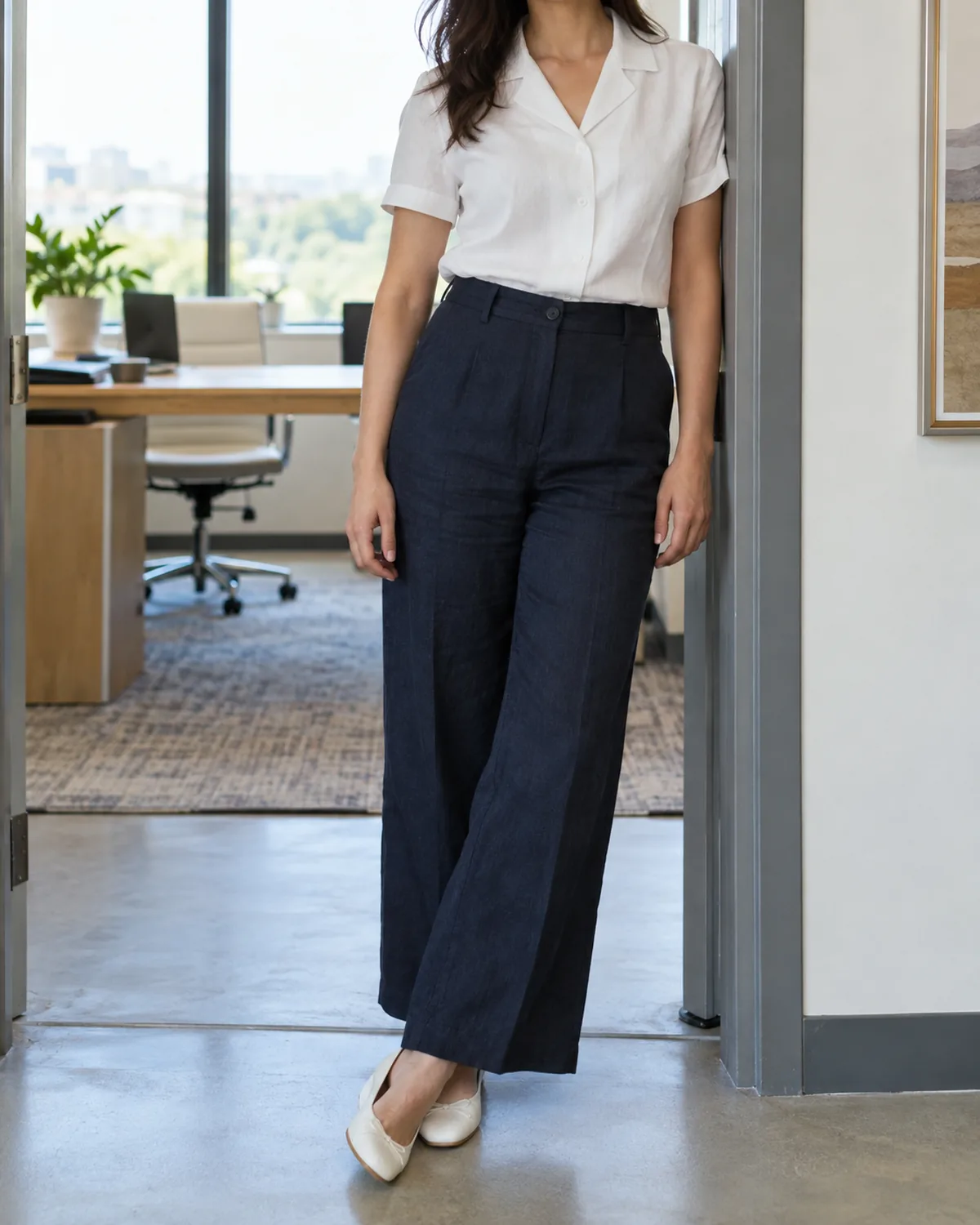 A woman standing in a doorway of a bright office wearing dark navy linen trousers, a white camp-collar blouse with short sleeves, and white leather flats