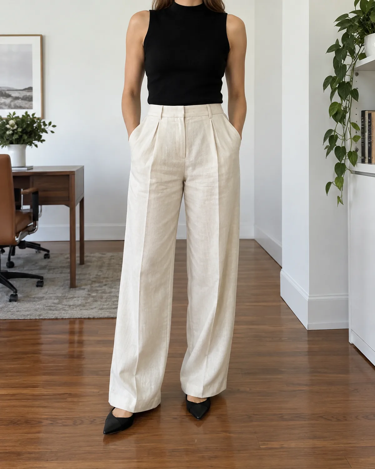 A woman standing in a bright office with both hands in the front pockets of cream pleated wide-leg linen trousers, wearing a black sleeveless mock-neck top and black pointed flats