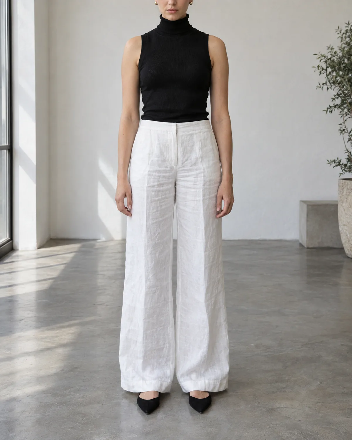 A woman standing full-length facing the camera in a bright space wearing white wide-leg linen trousers, a black sleeveless turtleneck, gold stud earrings, and black pointed flats