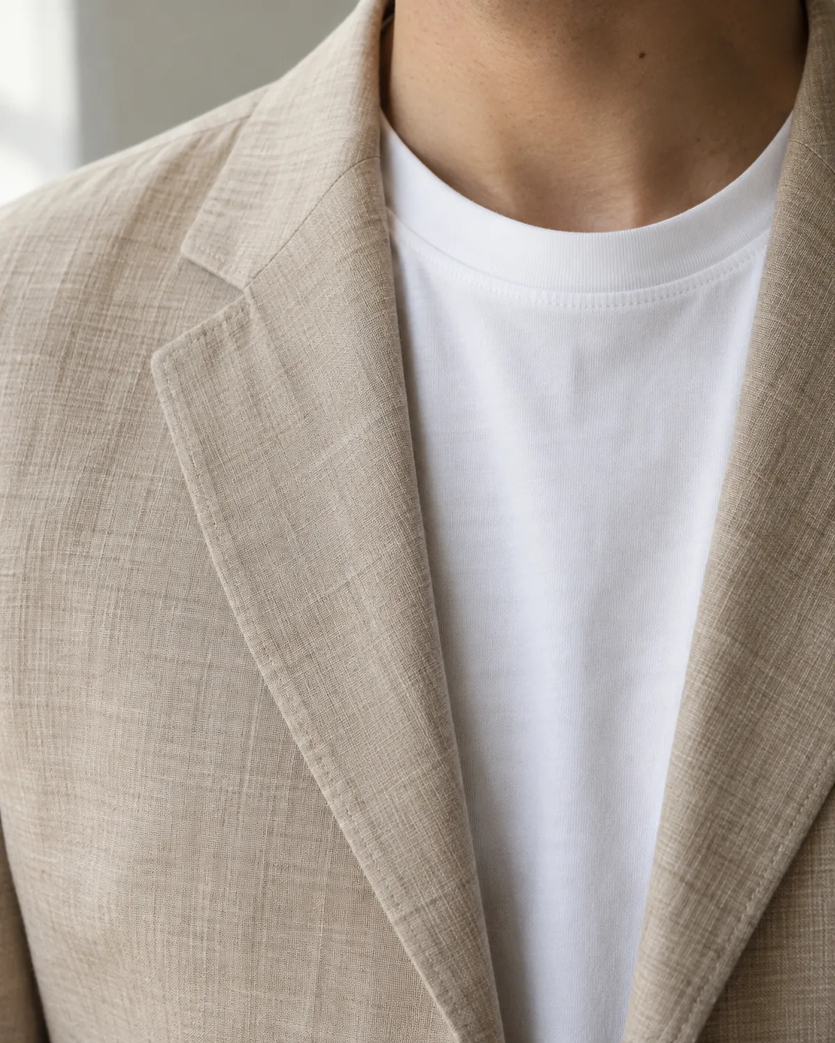 A close crop of a white cotton crew-neck tee visible under a sand linen blazer, showing the blazer lapel and the contrast between cotton knit and linen weave texture