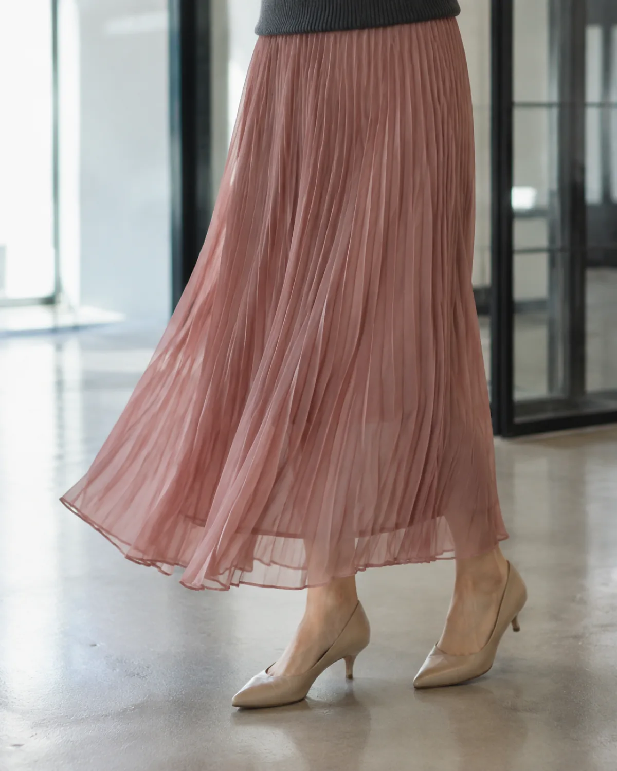 Close crop of a dusty rose pleated chiffon maxi skirt with accordion pleats and nude pointed kitten heels on a polished concrete floor