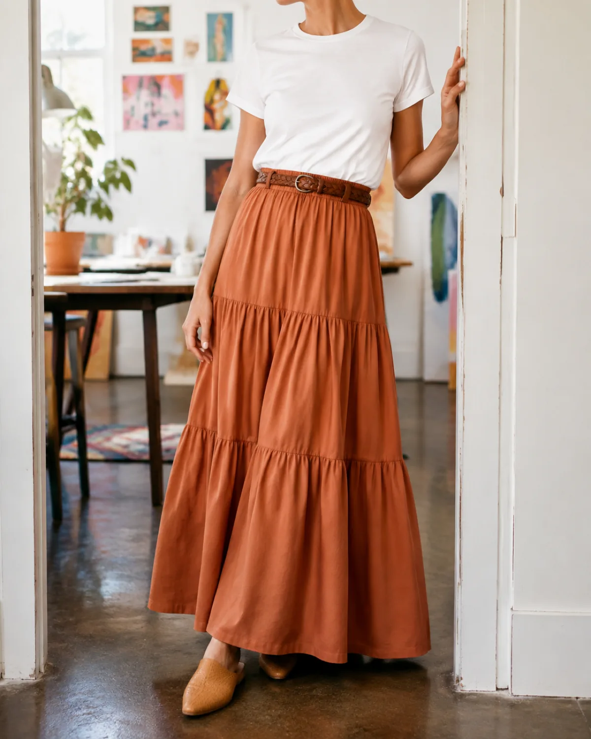 A woman in a terracotta tiered maxi skirt with a white structured tee and woven belt, leaning against a doorway in a creative studio