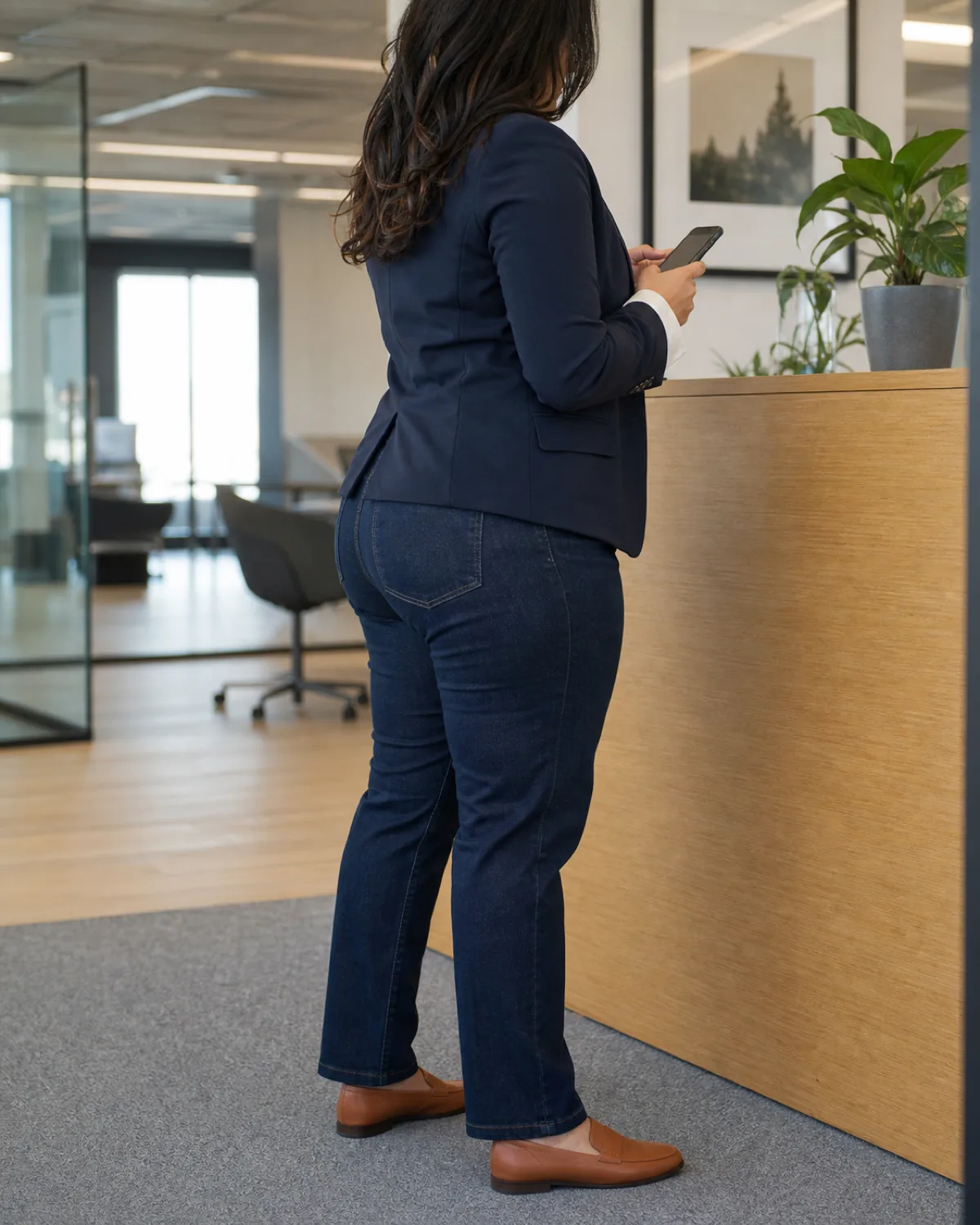 A midsize woman seen from behind in dark indigo straight-leg jeans, a white button-down shirt and a navy blazer, looking at a phone in an office corridor