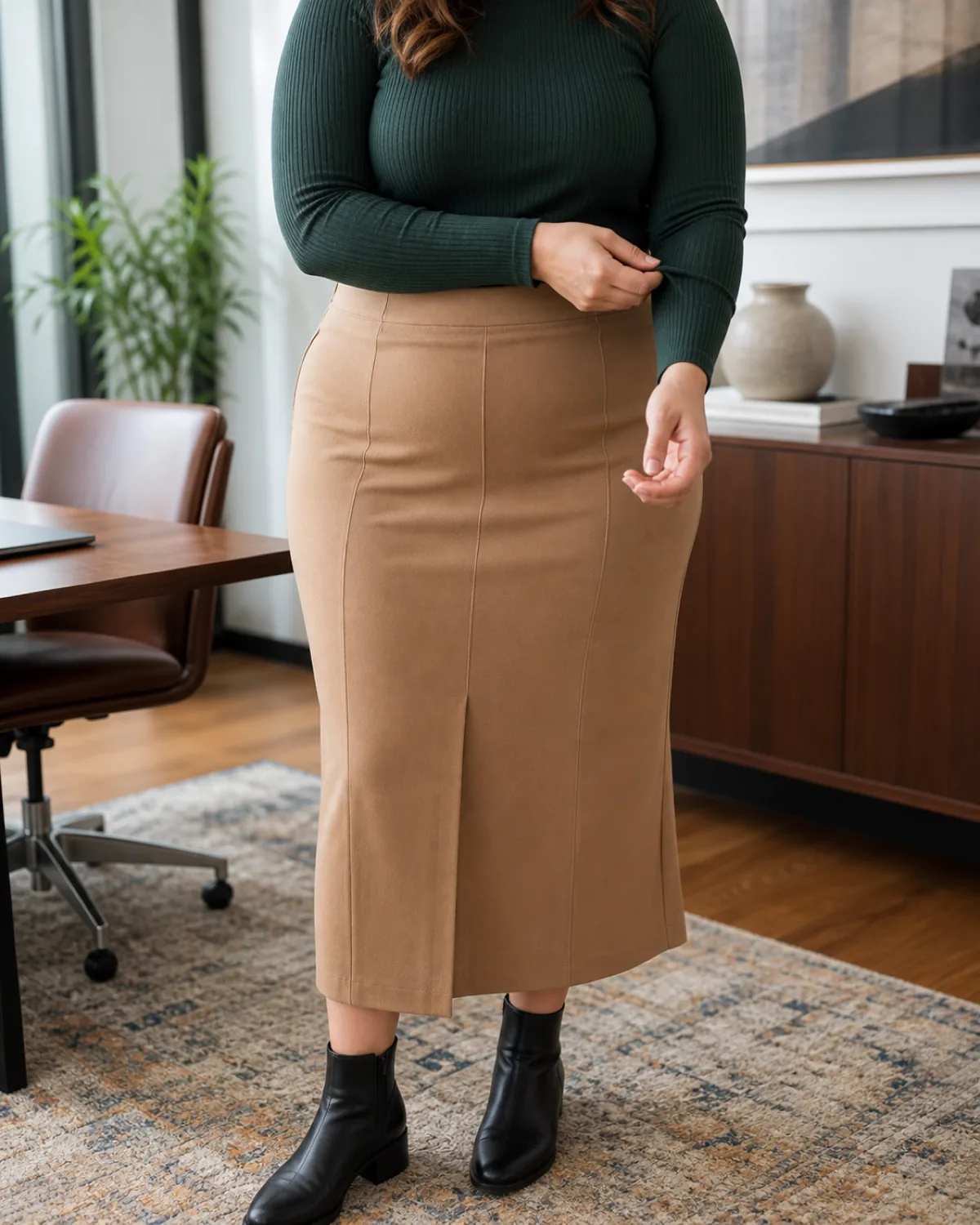 A midsize woman from chin to knee in a camel crepe midi skirt with a fitted dark green ribbed knit top tucked in, wearing black ankle boots, adjusting a sleeve cuff